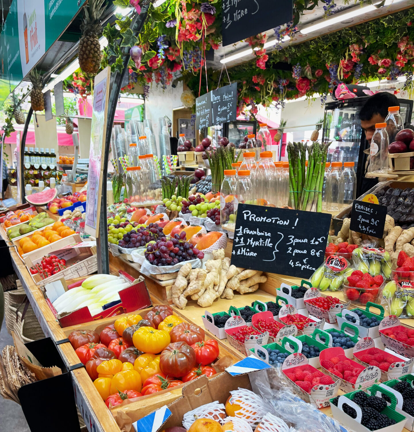 This image showcases a vibrant produce stand inside Marché Couvert des Enfants Rouges in Paris, filled with an array of fresh fruits and vegetables. The stall displays heirloom tomatoes, berries, grapes, ginger, asparagus, melons, and tropical fruits, along with fresh juices in bottles. Handwritten chalkboard signs indicate special promotions and pricing, while lush floral decorations hang from the ceiling, adding to the market’s charming and lively atmosphere. A feast for the eyes and a must-visit spot for food lovers!