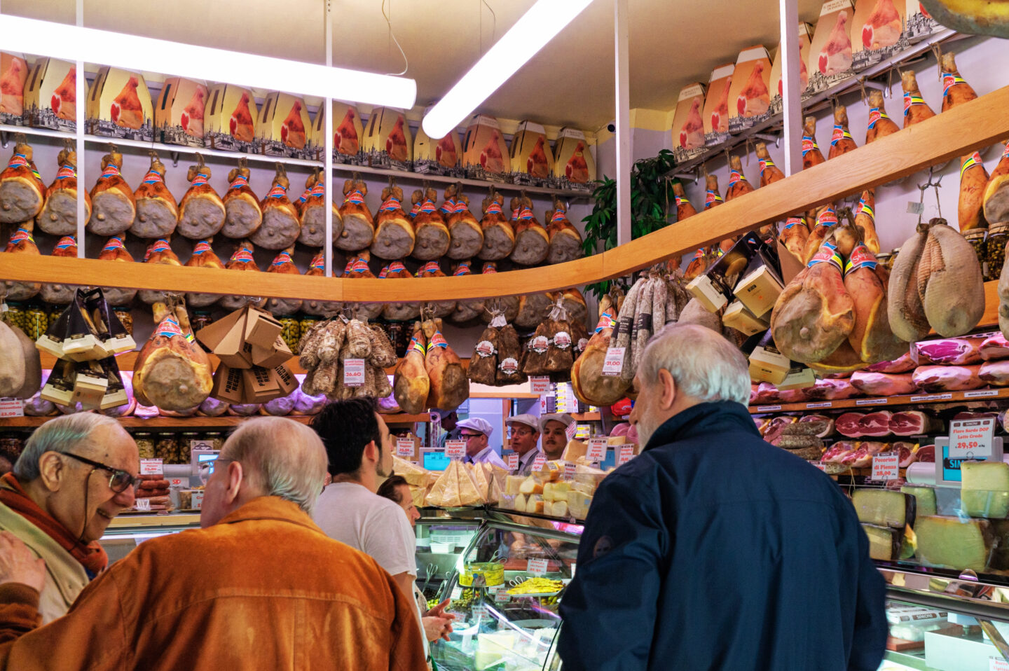 This image captures the vibrant interior of a traditional Italian deli inside Mercato di Mezzo in Bologna. The shop is lined with rows of hanging prosciutto, cured meats, and aged cheeses, creating an authentic and rustic atmosphere. Customers are engaged in conversation while butchers in white hats assist them from behind the glass display, which is filled with Parmigiano Reggiano, salami, and other gourmet specialties. A perfect snapshot of Bologna’s rich food culture!