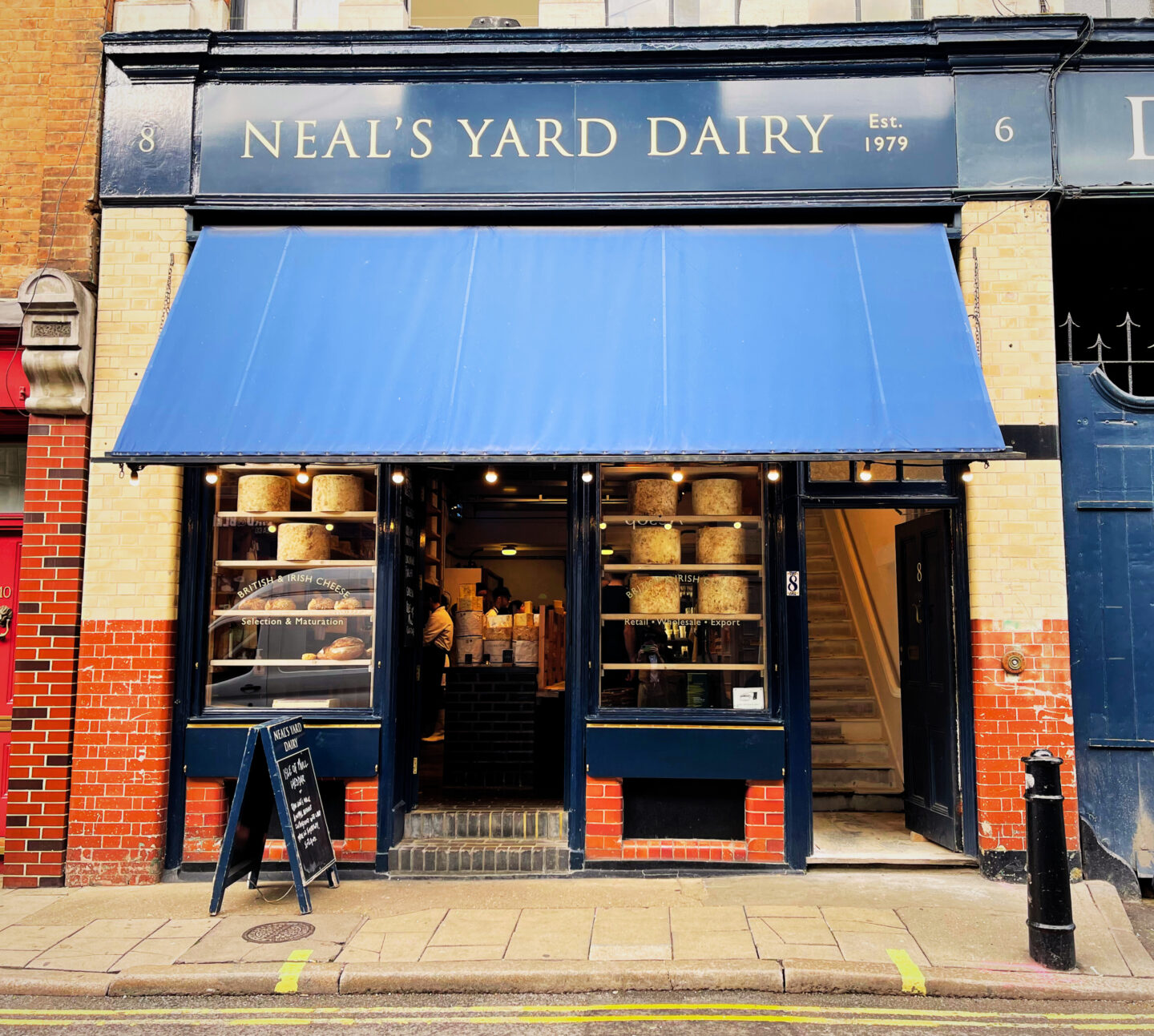 This image showcases the exterior of Neal’s Yard Dairy, a renowned cheese shop in London, established in 1979. The storefront, close to Borough Market in London, features a classic navy blue awning and signage, with large windows displaying impressive wheels of British and Irish cheeses stacked on wooden shelves. A small chalkboard sign stands outside, inviting customers in for a taste of artisanal dairy products. Its charming navy blue storefront, stacked with impressive wheels of cheese, makes it a recognisable and beloved destination for food lovers exploring the area. A must-visit if you're in Borough Market!
