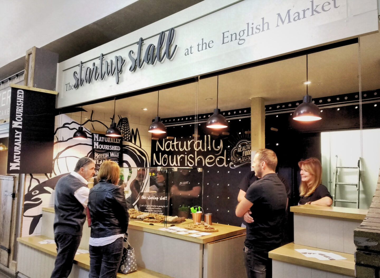 This image captures The Startup Stall at the English Market in Cork, Ireland, featuring Naturally Nourished, a vendor specialising in healthy, handmade food. The black and white signage highlights offerings like protein balls and plant-based snacks, while customers engage with the friendly staff behind the counter. Hanging industrial-style lights and a bold mural create a modern, inviting atmosphere in this historic market setting.