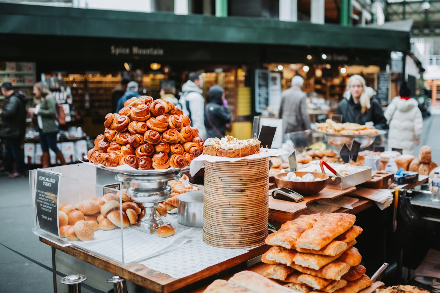 This image captures a bakery stall at Borough Market in London, showcasing a tempting display of freshly baked pastries, breads, and sweet treats. At the center, a towering stack of golden-brown cinnamon rolls sits atop a silver platter, surrounded by loaves of focaccia, buns, and slices of cake. In the background, market-goers browse stalls filled with gourmet ingredients, adding to the lively and bustling atmosphere of this iconic food market.