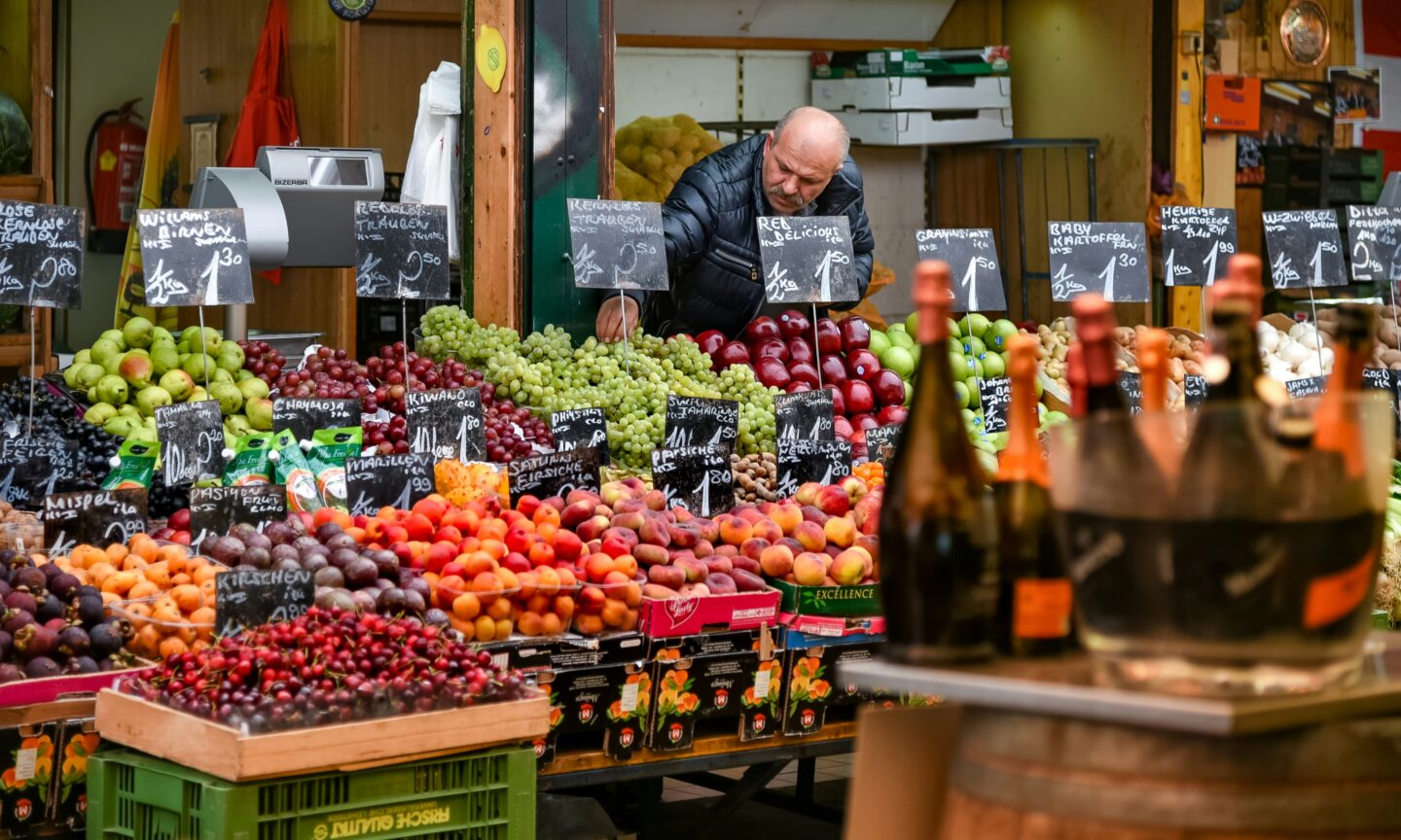 This image captures a fruit and vegetable stand at Naschmarkt in Vienna, Austria, a famous open-air market known for its diverse selection of fresh produce, international foods, and specialty goods. A vendor arranges grapes, apples, pears, cherries, peaches, and exotic fruits, all neatly displayed with handwritten price tags on chalkboards. In the foreground, bottles of sparkling wine chill in an ice bucket, adding to the lively and inviting atmosphere of this iconic market.