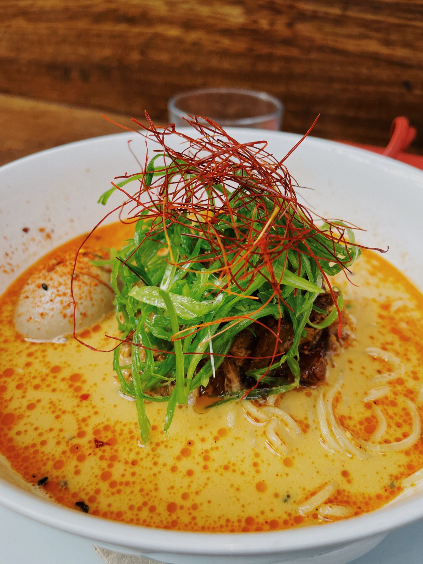 Close-up of a vibrant ramen bowl with creamy orange broth, sliced green onions, a soft-boiled egg, chili oil, and crispy toppings served at Mackie Mayor.