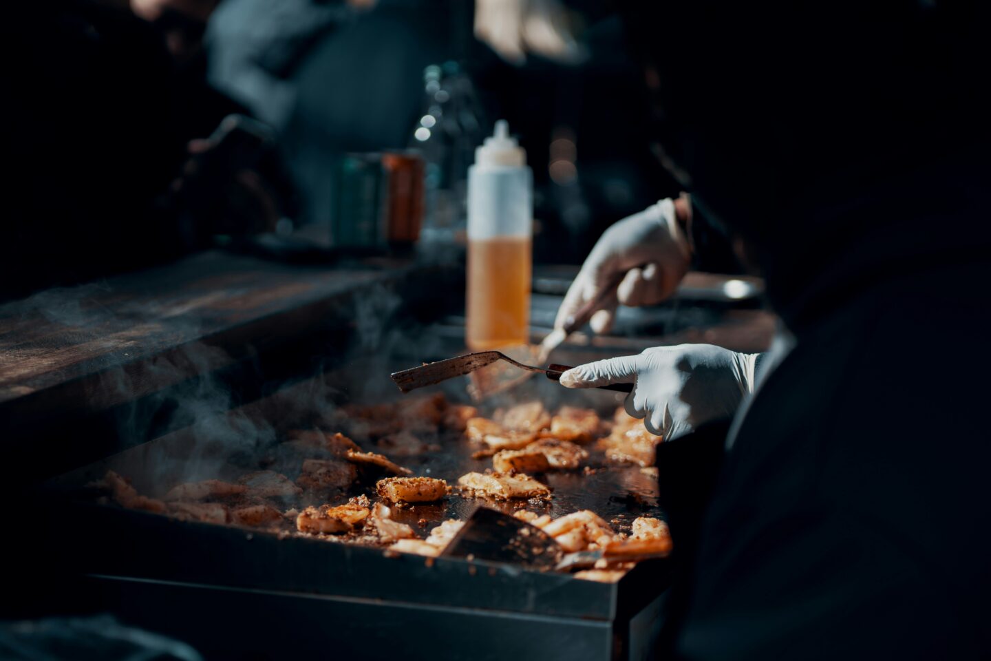 Close-up of a gloved street food vendor cooking seasoned food on a hot griddle, with steam rising and a bottle of oil blurred in the background.