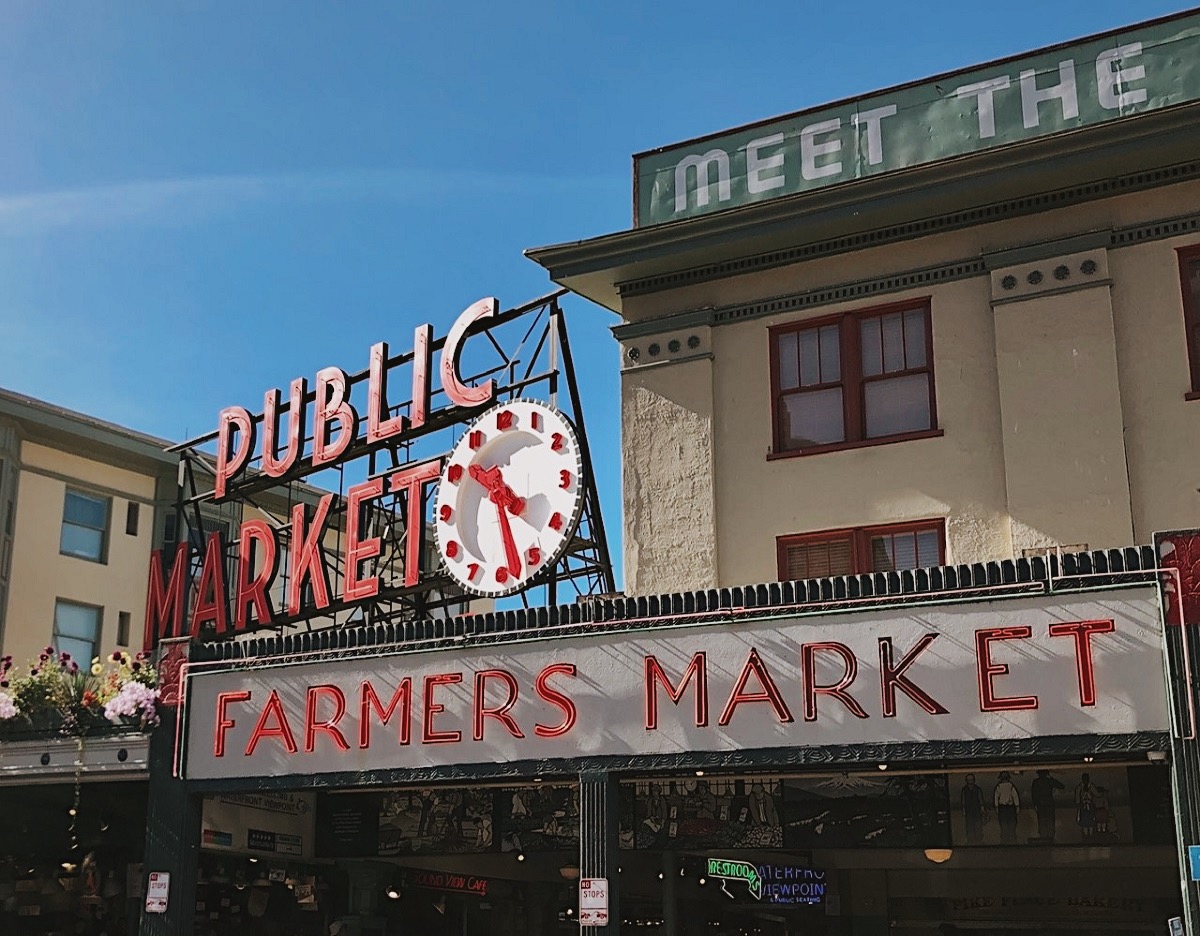 Daytime shot of Seattle’s Pike Place Market featuring the iconic “Public Market” red neon sign with a clock showing 11:55, and the “Farmers Market” sign below. A vintage green sign above reads “Meet the Producer,” and hanging flower baskets line the building’s edge.