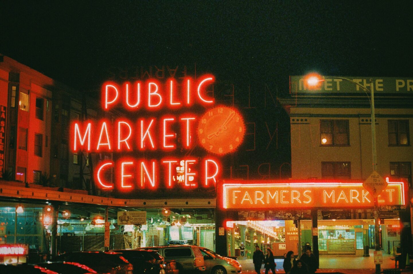 Nighttime photo of the iconic neon signs at Seattle’s Pike Place Market, featuring the bright red “Public Market Center” sign with a clock in the middle, and the adjacent glowing “Farmers Market” sign below. The surrounding area is lit with storefront lights and a few people are walking under the signage.      