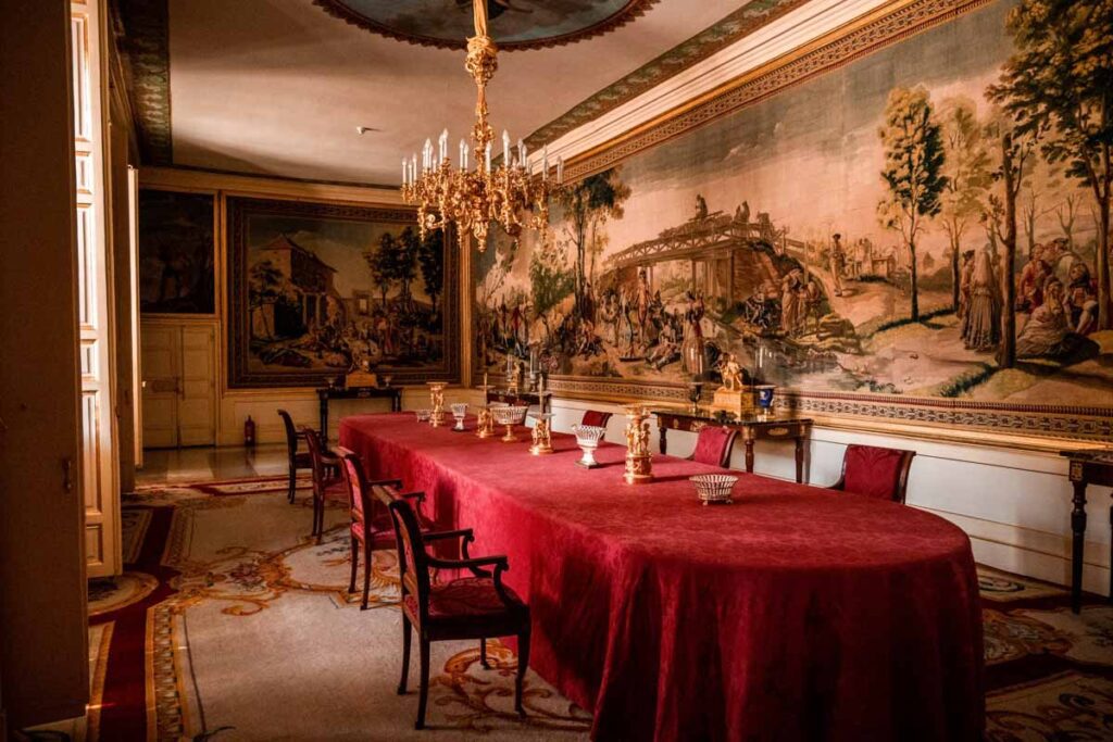 San Lorenzo de El Escorial: Lavish dining room with a long red-covered table, gold chandeliers, and large detailed tapestries depicting pastoral scenes on the walls.