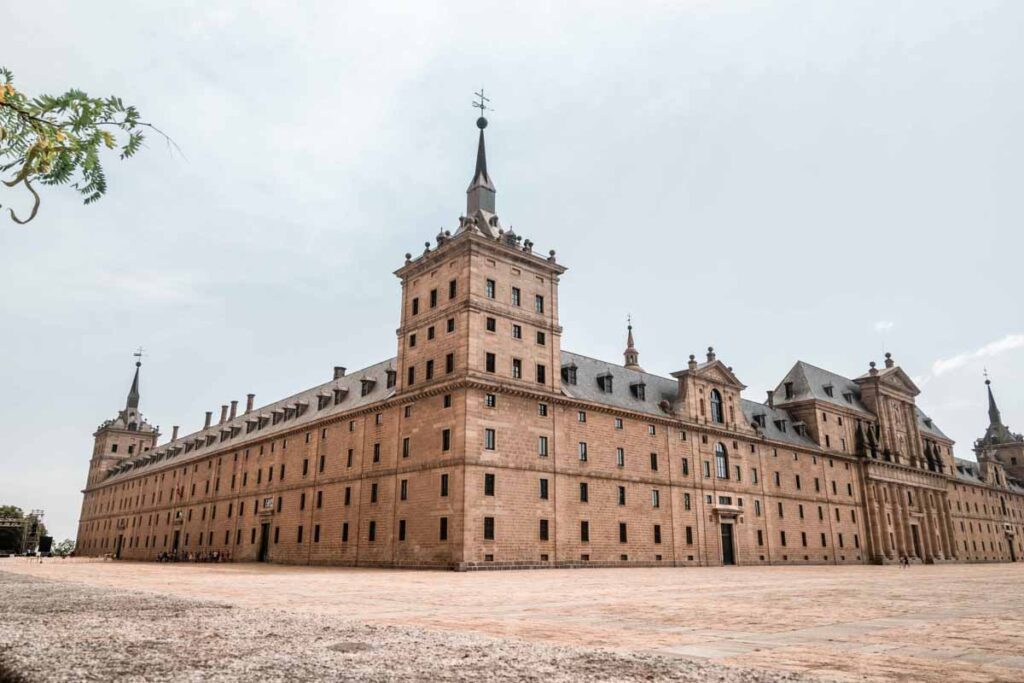 Exterior of El Escorial Monastery: Wide-angle view of the grand façade of El Escorial, showing its imposing rectangular architecture, towers with spires, and stone courtyard.