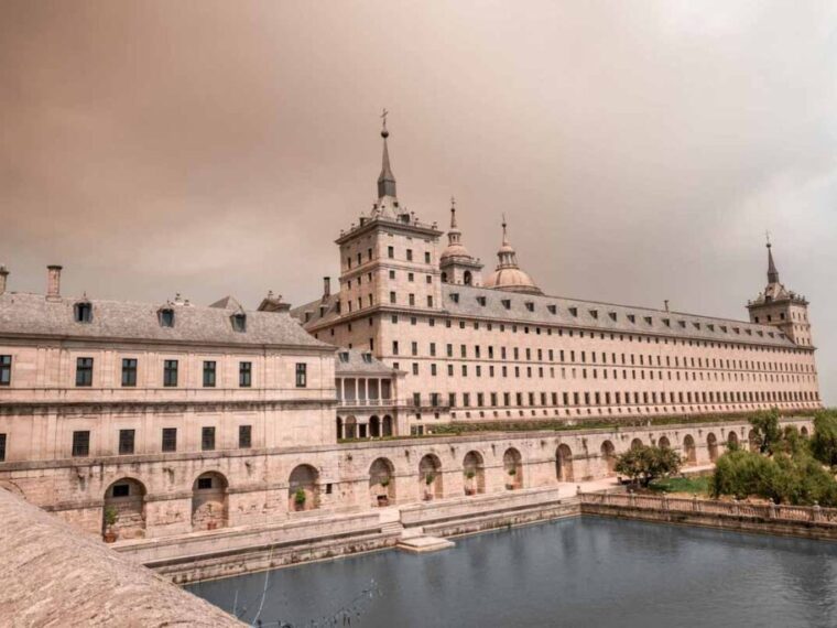 View of the massive El Escorial complex with its towers and domes rising above a reflecting pool and a terraced garden wall.