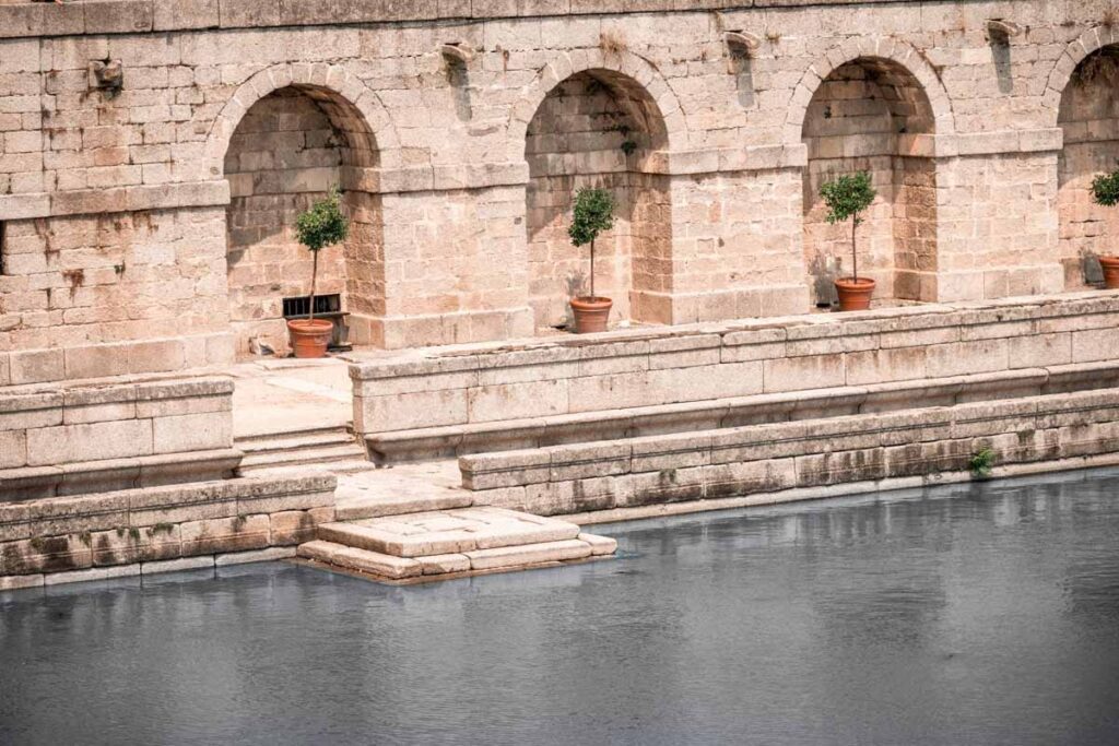 Close-up of a stone terrace with arched alcoves and terracotta pots holding small trees, lining the edge of a peaceful reflecting pool in San Lorenzo de El Escorial