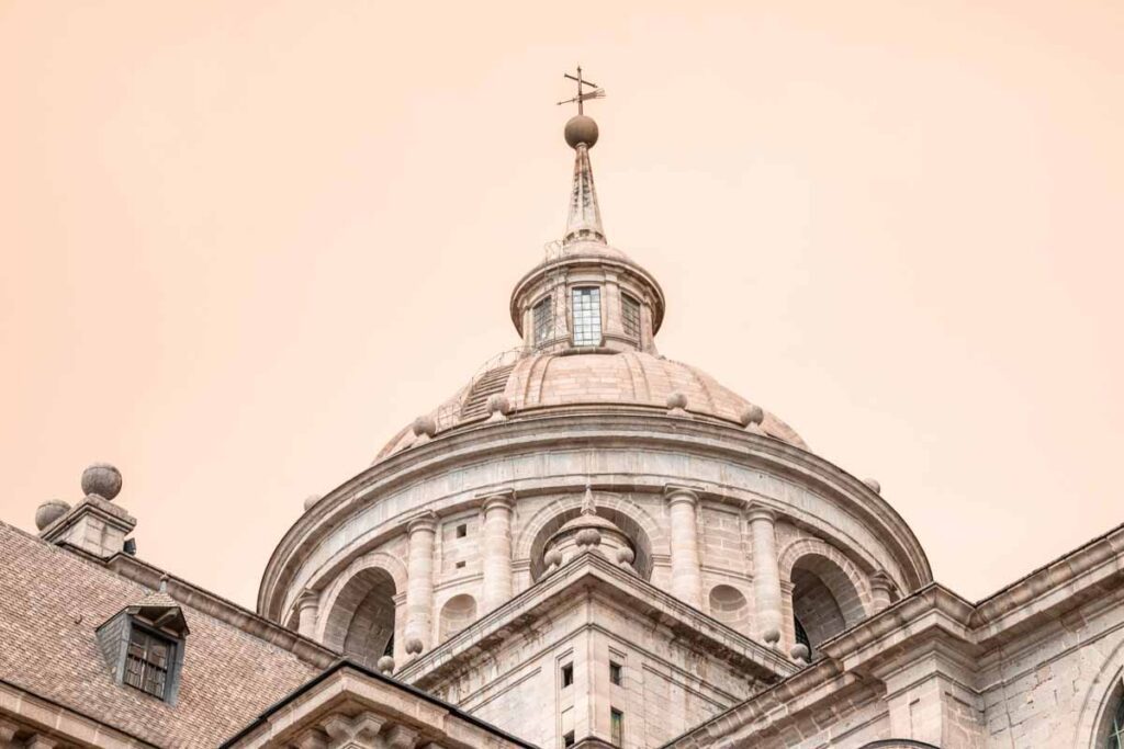 Close-Up of El Escorial Dome: Upward view of the dome of El Escorial with its arched windows, stone columns, and a cross-topped spire set against a soft peach sky.