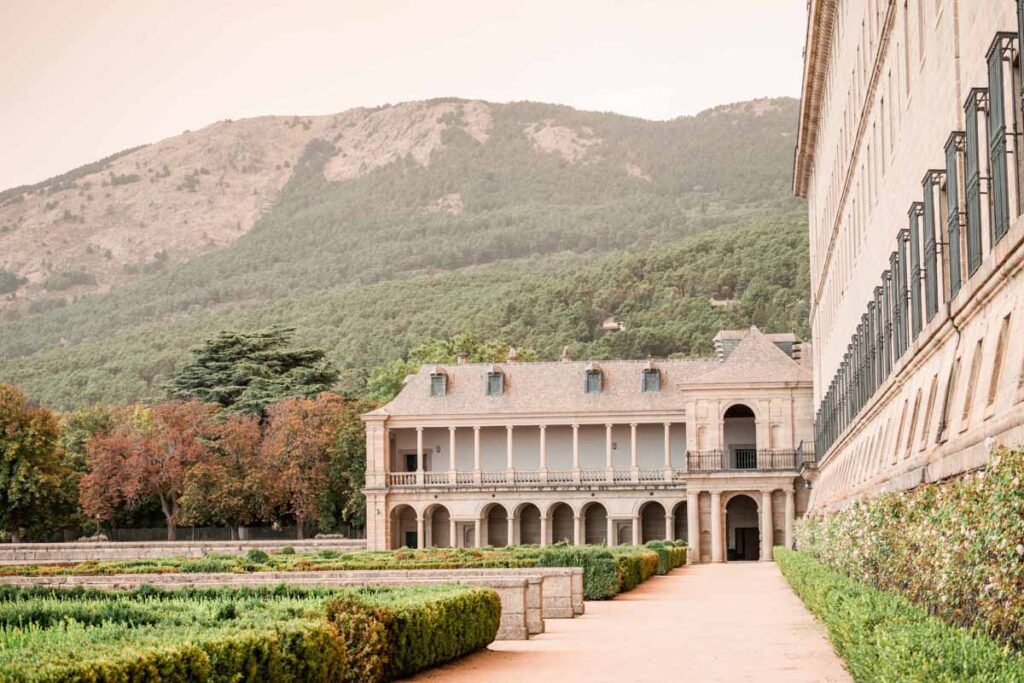 Scenic view of manicured gardens and a two-story stone building with arched walkways, set against a forested mountain backdrop beside El Escorial.