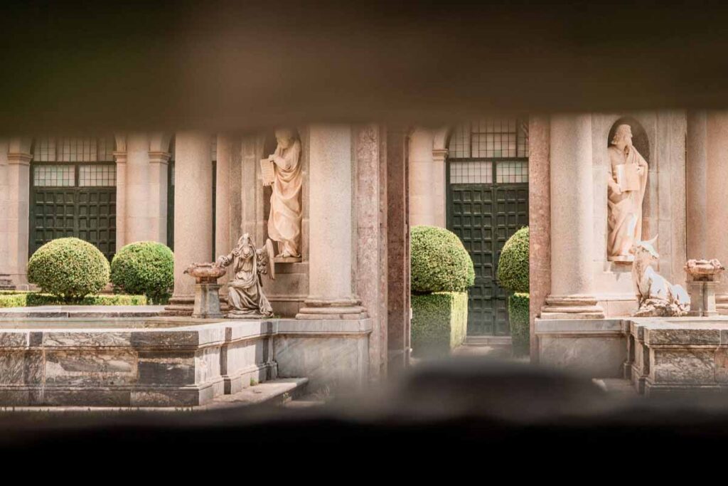 San Lorenzo de El Escorial: View of a stone courtyard framed by archways, with classical statues, manicured spherical hedges, and a marble fountain, seen through a dark window or gate.