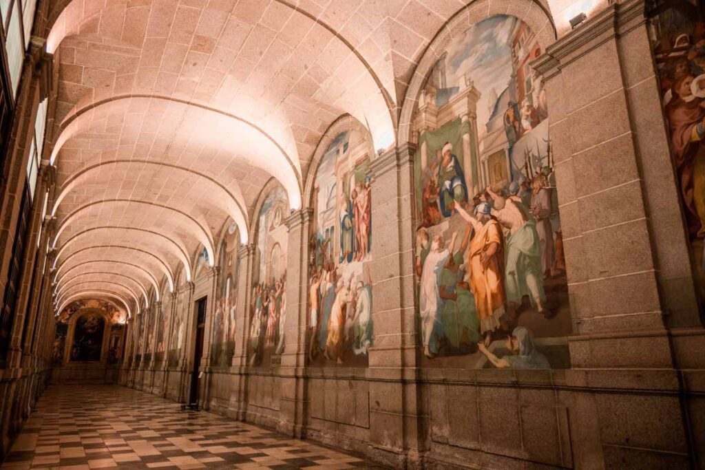 Ornate corridor inside San Lorenzo de El Escorial featuring arched stone ceilings and colorful frescoes depicting religious scenes along the walls.
