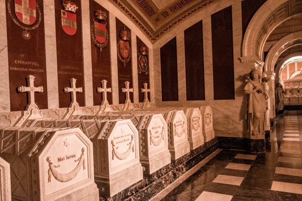 San Lorenzo de El Escorial: Interior of the Royal Crypt with intricately carved marble tombs, gold-accented crests, and stone walls engraved with names of Spanish royalty.