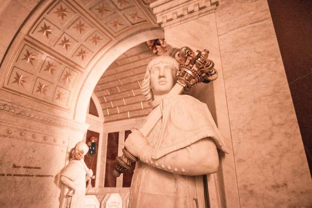 San Lorenzo de El Escorial: Close-up of a white marble statue of a royal figure holding an ornate golden scepter, under a vaulted ceiling decorated with gold stars.