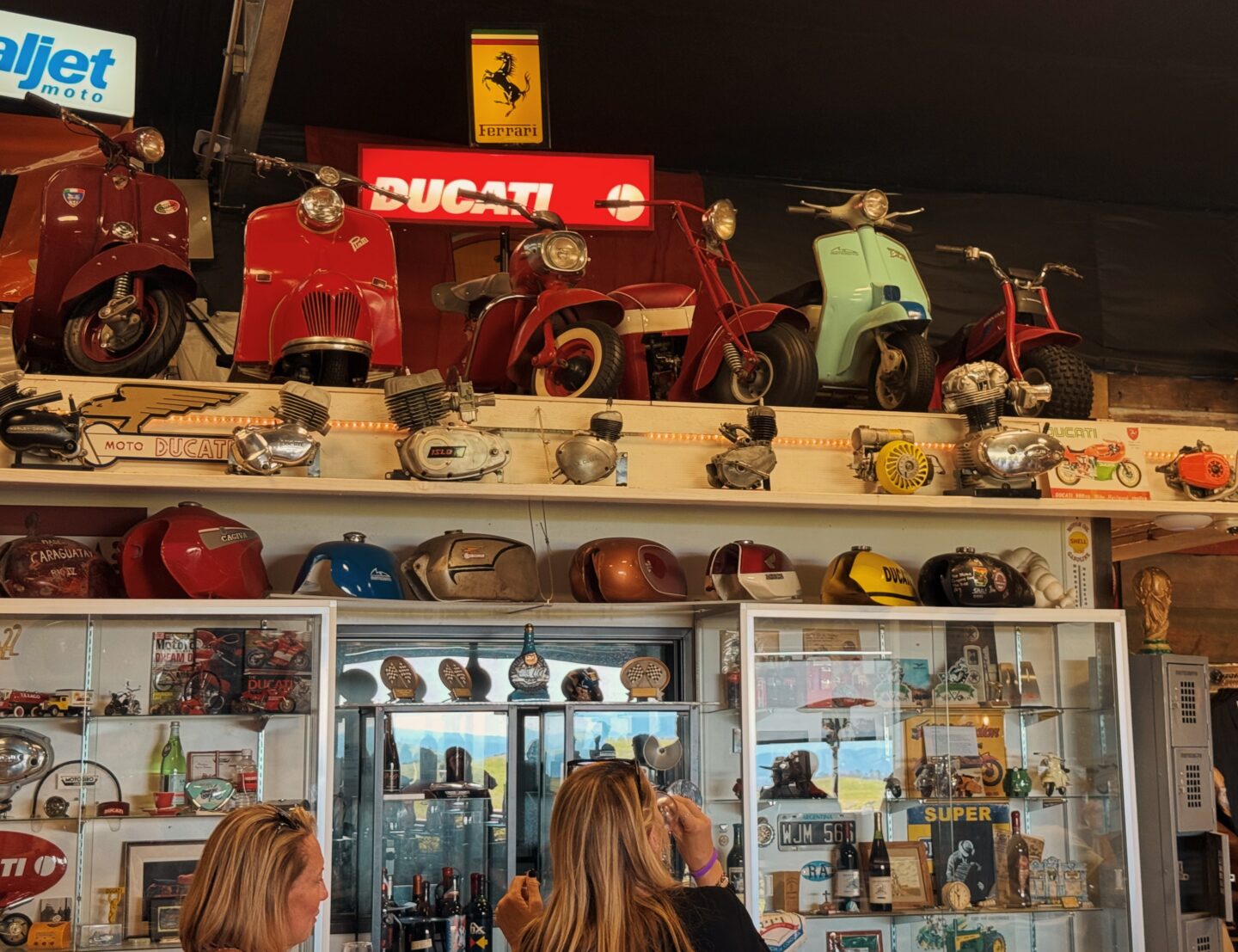 Vintage scooters and motorcycles displayed above glass cases inside Doffo Winery in Temecula in winter with a bright red "Ducati" sign and a yellow "Ferrari" logo on the wall. Visitors stand below viewing collections of helmets, engine parts, and memorabilia in a lively tasting room setting.