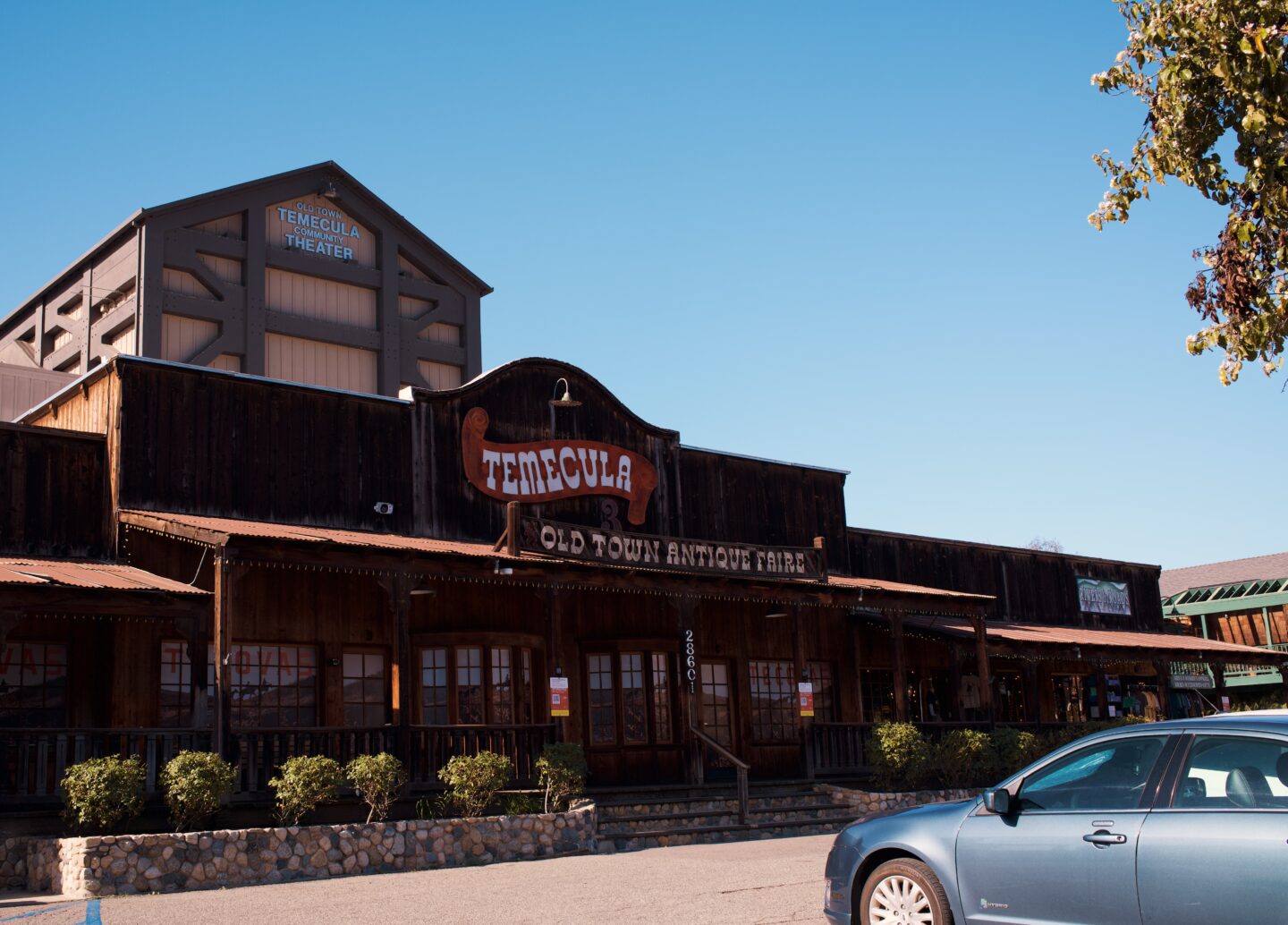 ustic wooden storefront in Old Town Temecula in winter with a large sign reading "Temecula Old Town Antique Faire" across the front. The Old Town Temecula Community Theater rises behind the building under a clear blue sky with cars parked out front.