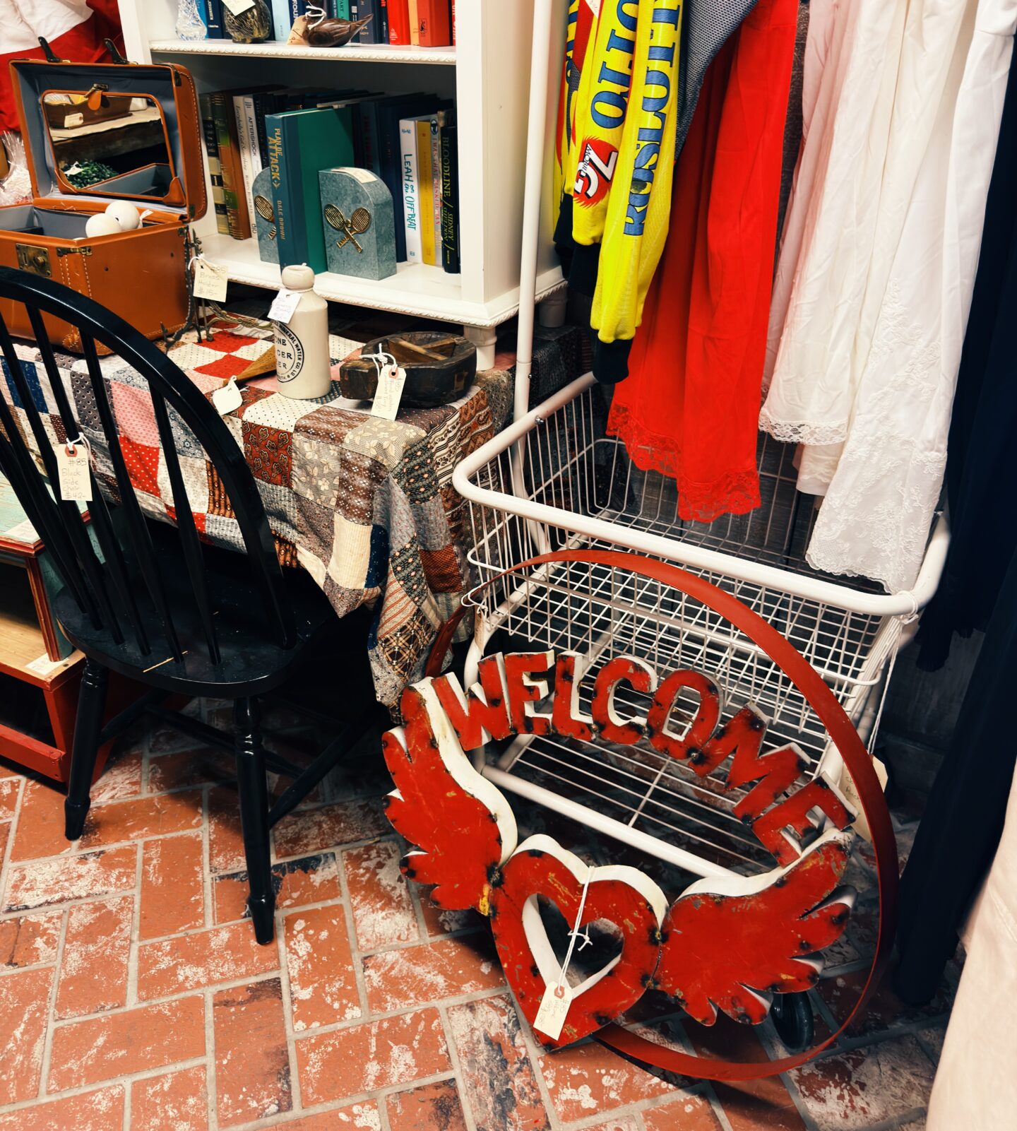 Vintage shop display in Old Town Temecula in winter featuring clothing on a rack, books on shelves, and small antiques arranged on a table. A red sign on the floor reads "WELCOME" with a heart and wings, adding a cozy and eclectic shopping atmosphere.