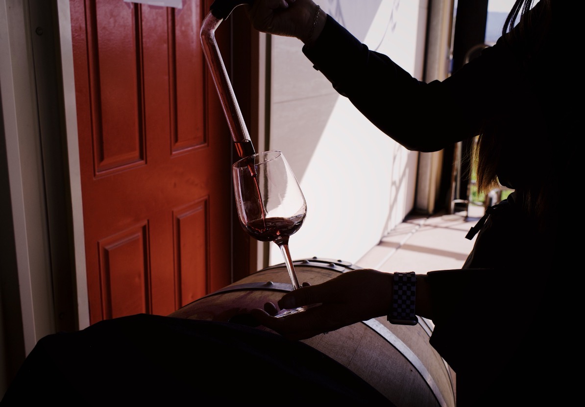 Red wine being poured into a glass from a tasting tube over a wooden barrel on the patio at Doffo Winery in Temecula in winter. Natural sunlight and open air create a relaxed outdoor tasting experience.