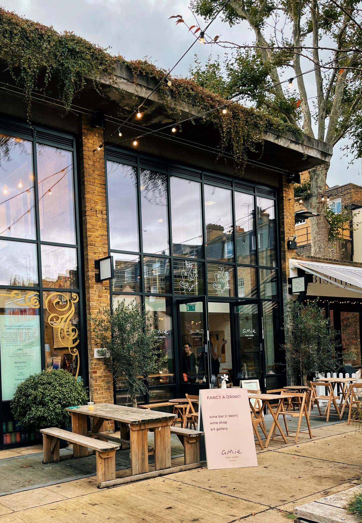 Amie Wine Studio with large glass windows outdoor wooden tables and string lights surrounded by greenery. A pink sign outside reads "FANCY A DRINK. wine bar plus beer. wine shop. art gallery. amie wine studio."