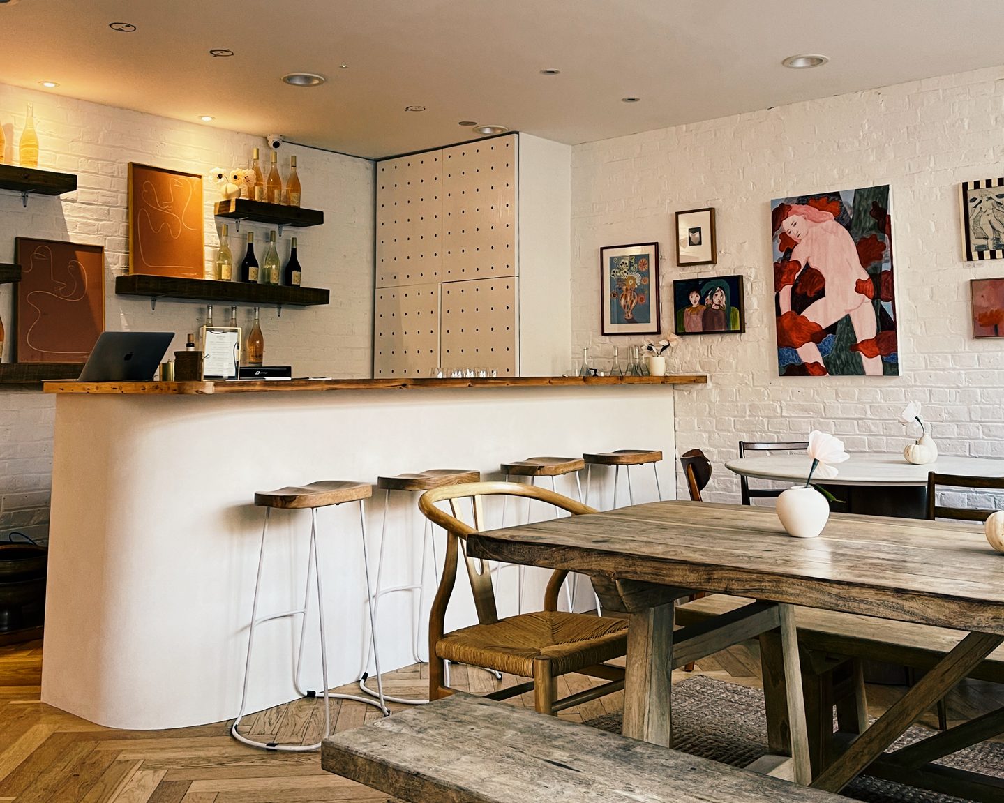 Interior of Amie Wine Studio with a white curved bar rustic wooden tables and wine bottles displayed on dark shelves. Colorful artwork hangs across the white brick walls giving the London wine bar the feel of a relaxed art gallery.