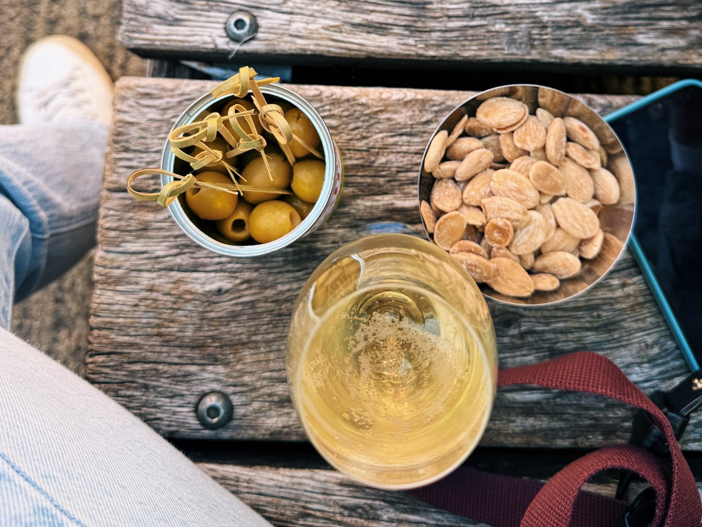 Glass of white wine with bowls of green olives and salted almonds on a weathered wooden table at Amie Wine Studio. The simple wine and snack pairing is served outside the London wine bar.