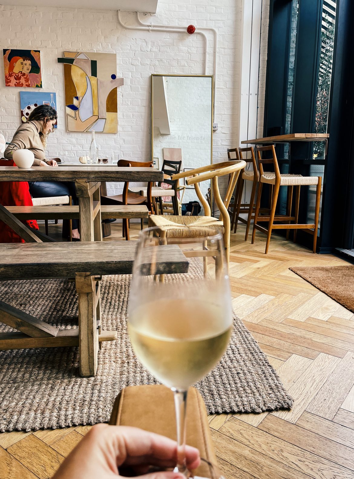 Glass of white wine held in the foreground inside Amie Wine Studio with rustic wooden tables woven chairs and colorful artwork behind it. The bright relaxed interior combines a London wine bar with an art gallery.