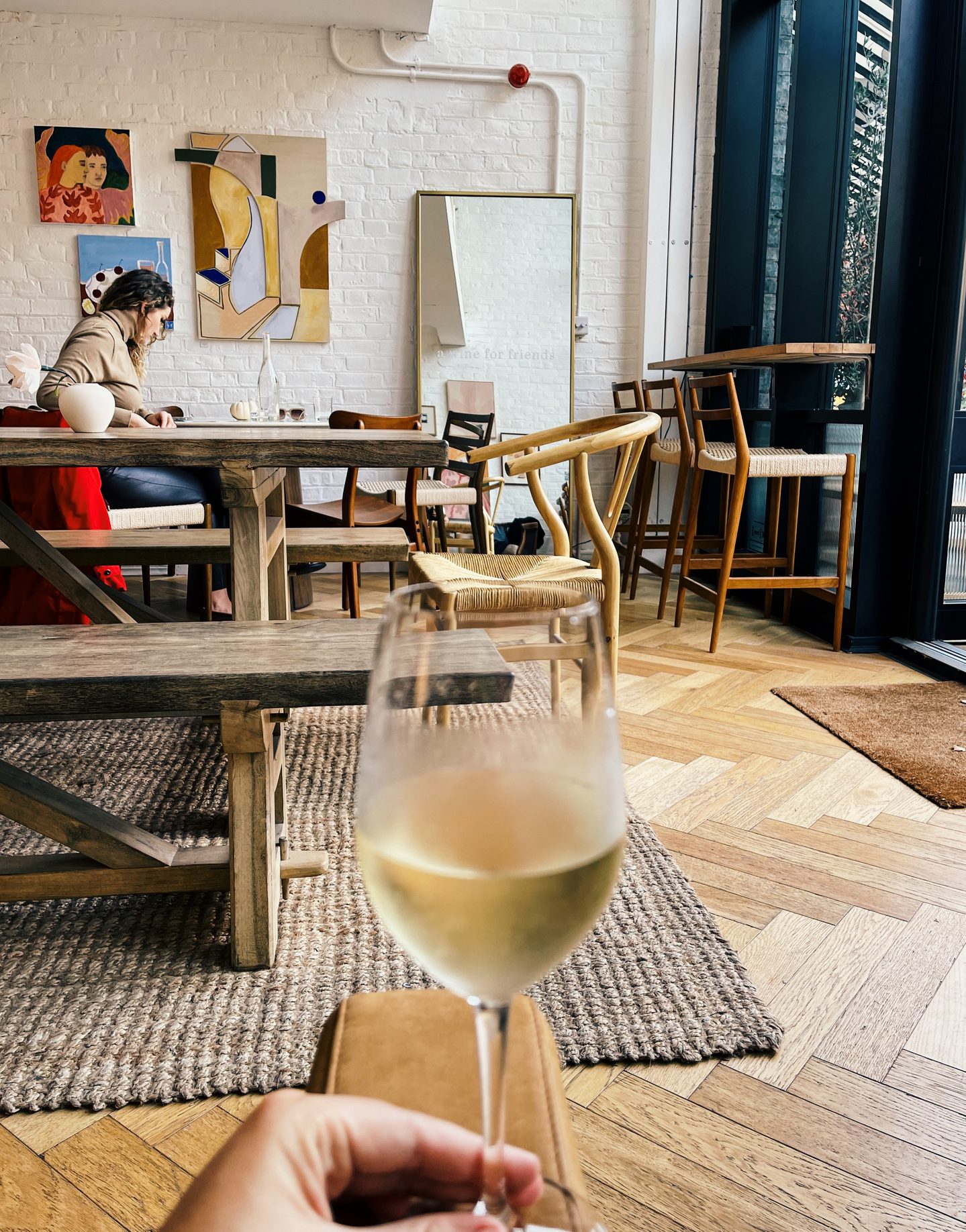 Glass of white wine held in the foreground inside Amie Wine Studio with rustic wooden tables woven chairs and colorful artwork behind it. The bright relaxed interior combines a London wine bar with an art gallery.