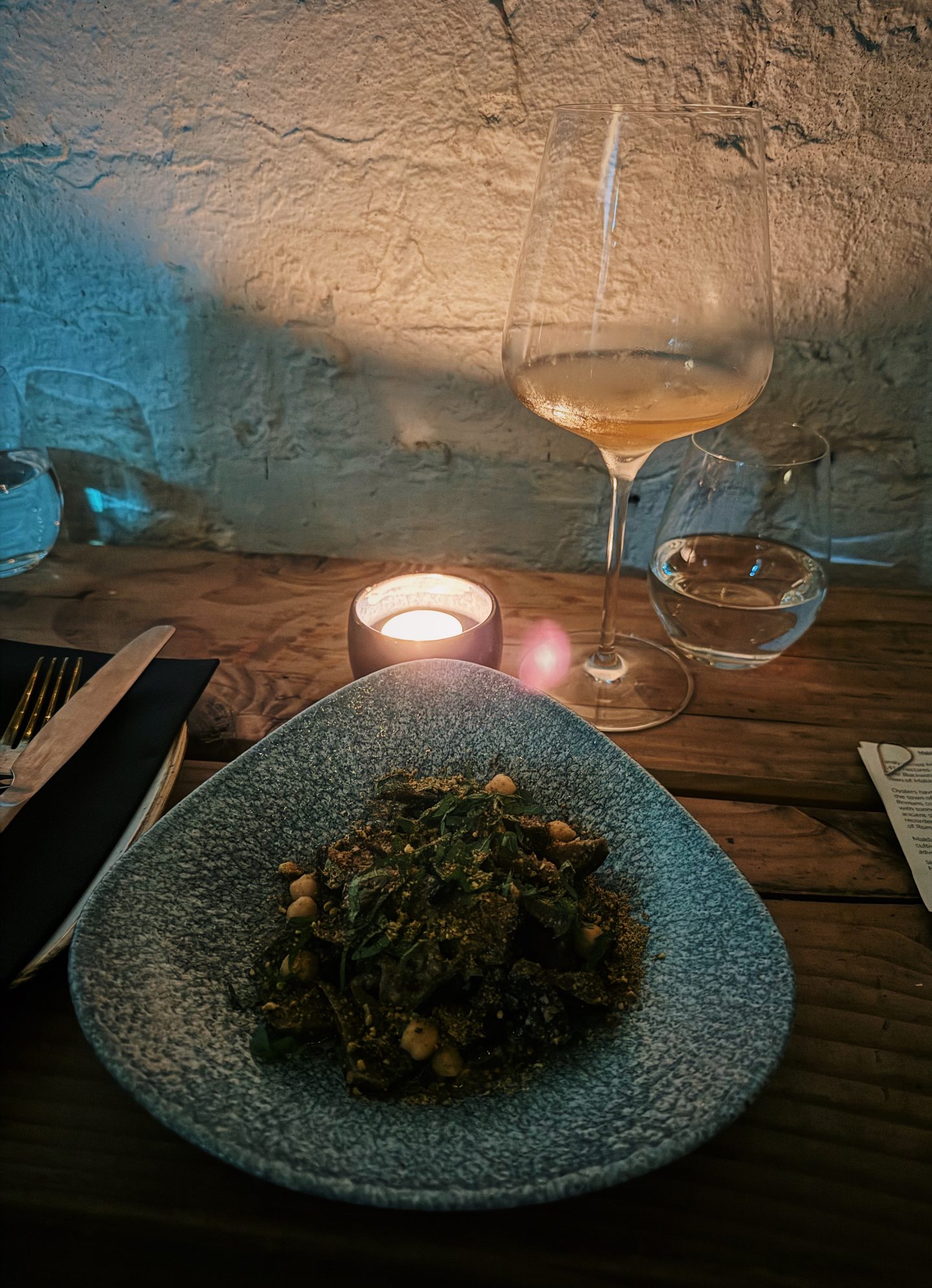 Candlelit dinner at Aspen & Meursault with a textured blue bowl of greens and chickpeas beside a glass of pale wine. Warm candlelight and a rustic stone wall create an intimate wine bar atmosphere.