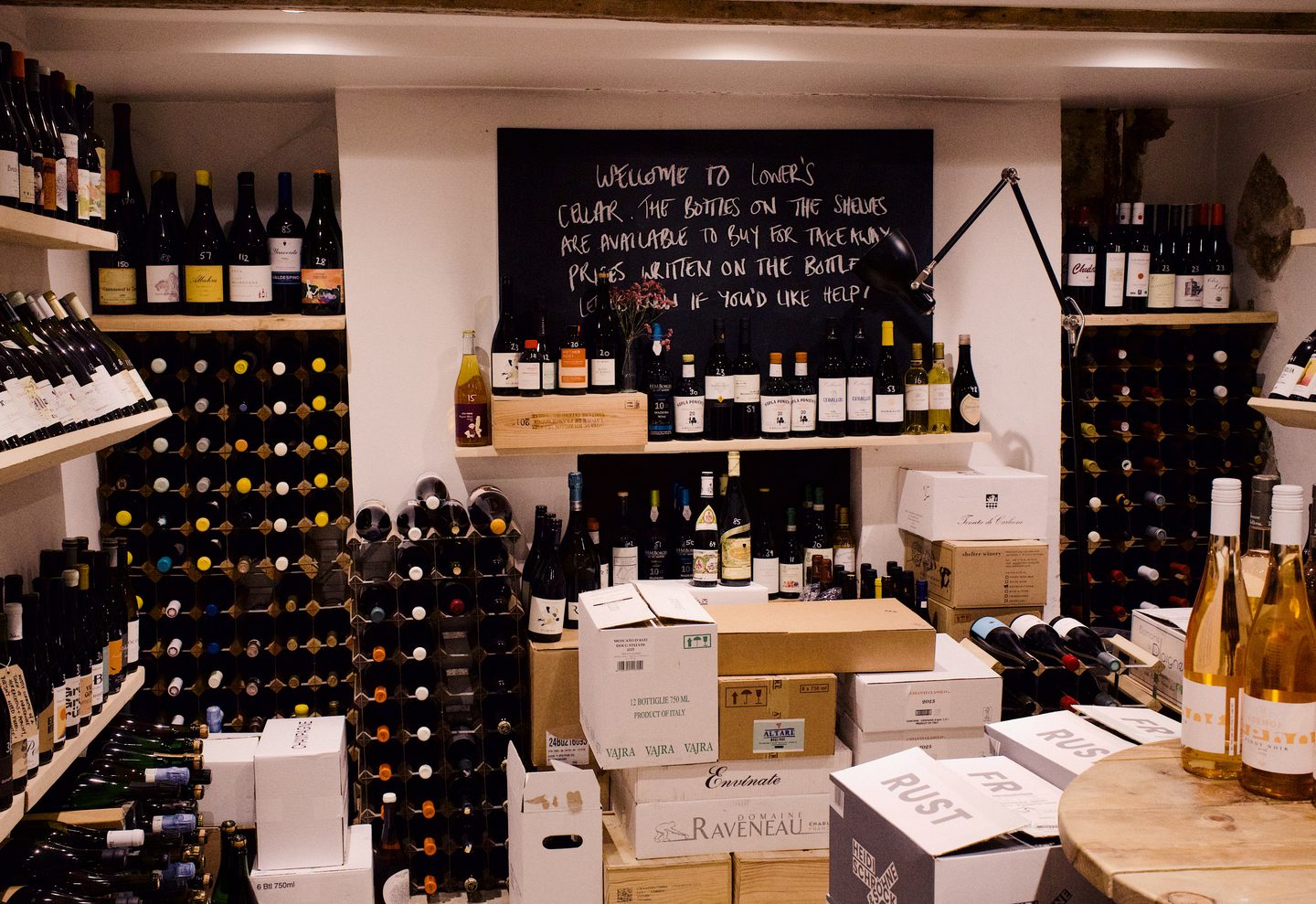 Floor to ceiling racks of wine bottles and stacked cases fill the cellar at Lower Wine Bar in Waterloo. A handwritten chalkboard welcomes visitors to the cellar and explains that bottles on the shelves are available to buy at takeaway prices.