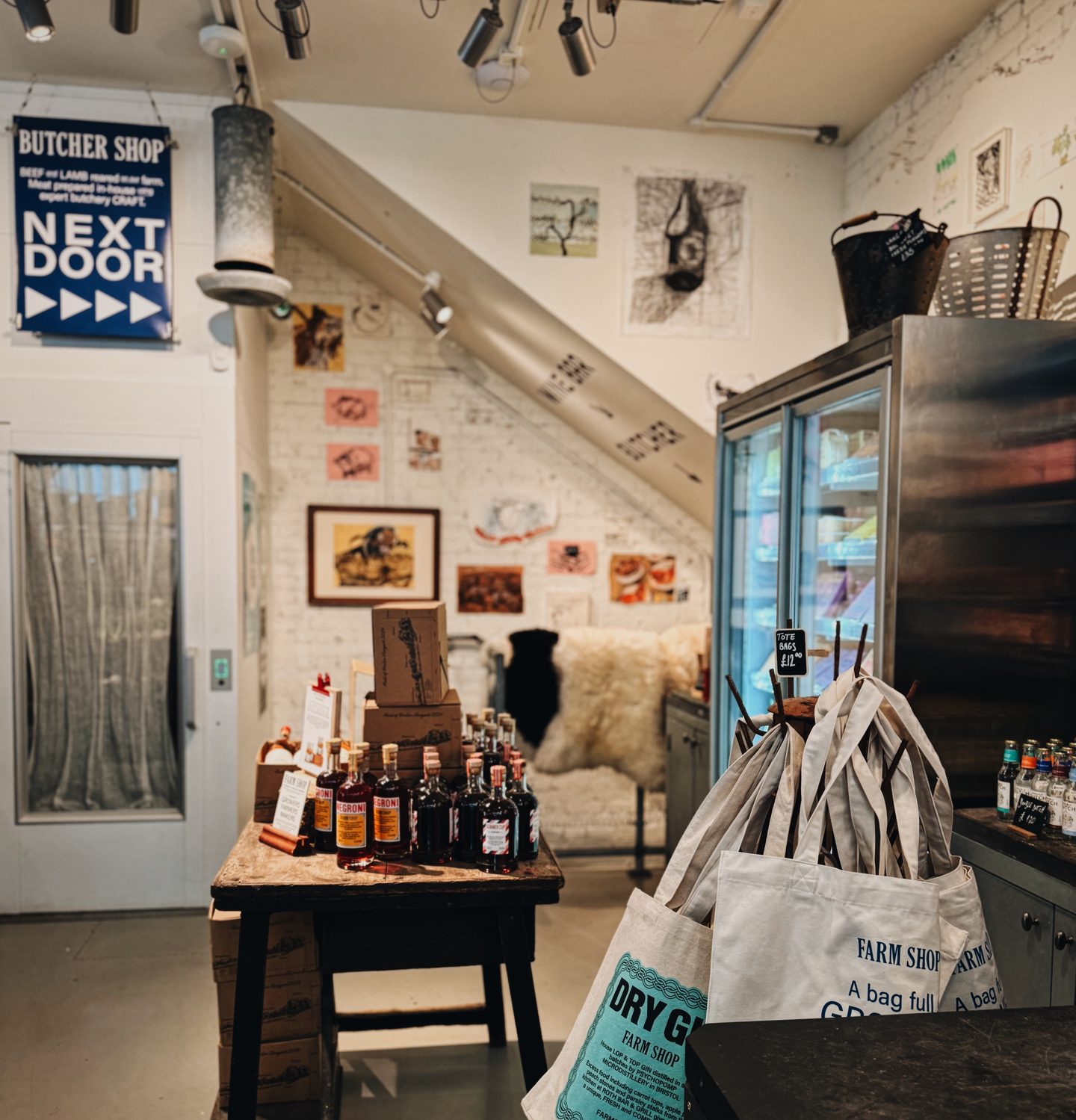 Upstairs shop at Farm Shop Mayfair with bottles displayed on a rustic wooden table and tote bags beside a refrigerated case. Signs reading "BUTCHER SHOP NEXT DOOR" and "FARM SHOP" add to the relaxed market style interior.