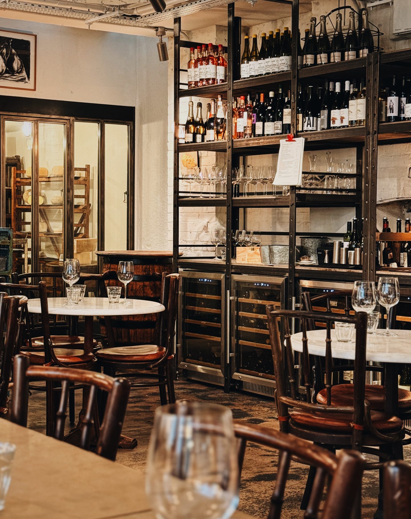 Downstairs wine bar at Farm Shop Mayfair with marble tables dark wooden chairs and tall shelves filled with wine bottles and glasses. Wine fridges and a glass fronted cheese room give the intimate space the feel of a traditional London wine bar.