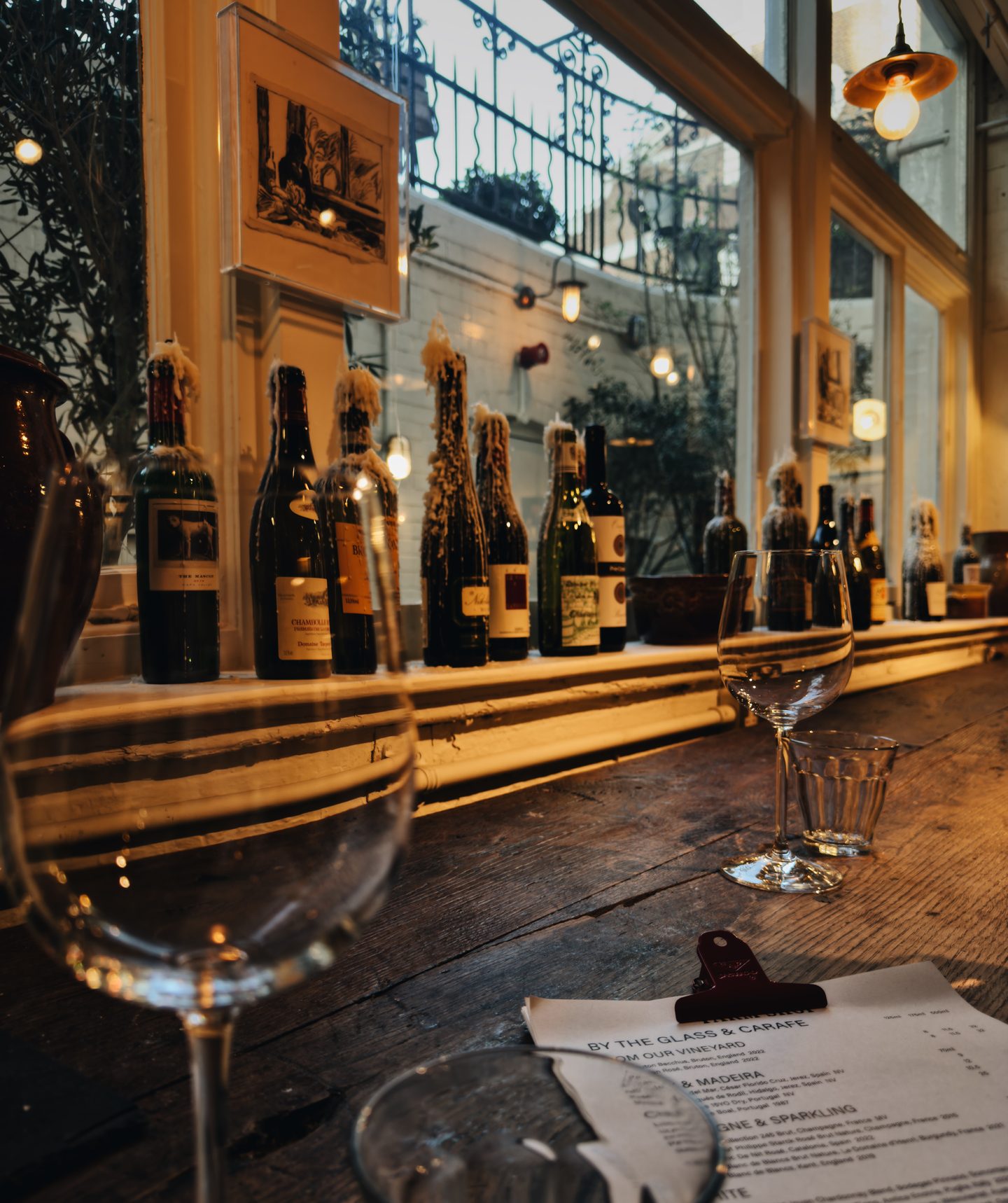 Wine bottles with dripped wax seals line the window beside wooden counter seating at the downstairs wine bar at Farm Shop Mayfair. Empty wine glasses and warm pendant lights create a cozy setting overlooking a small courtyard.