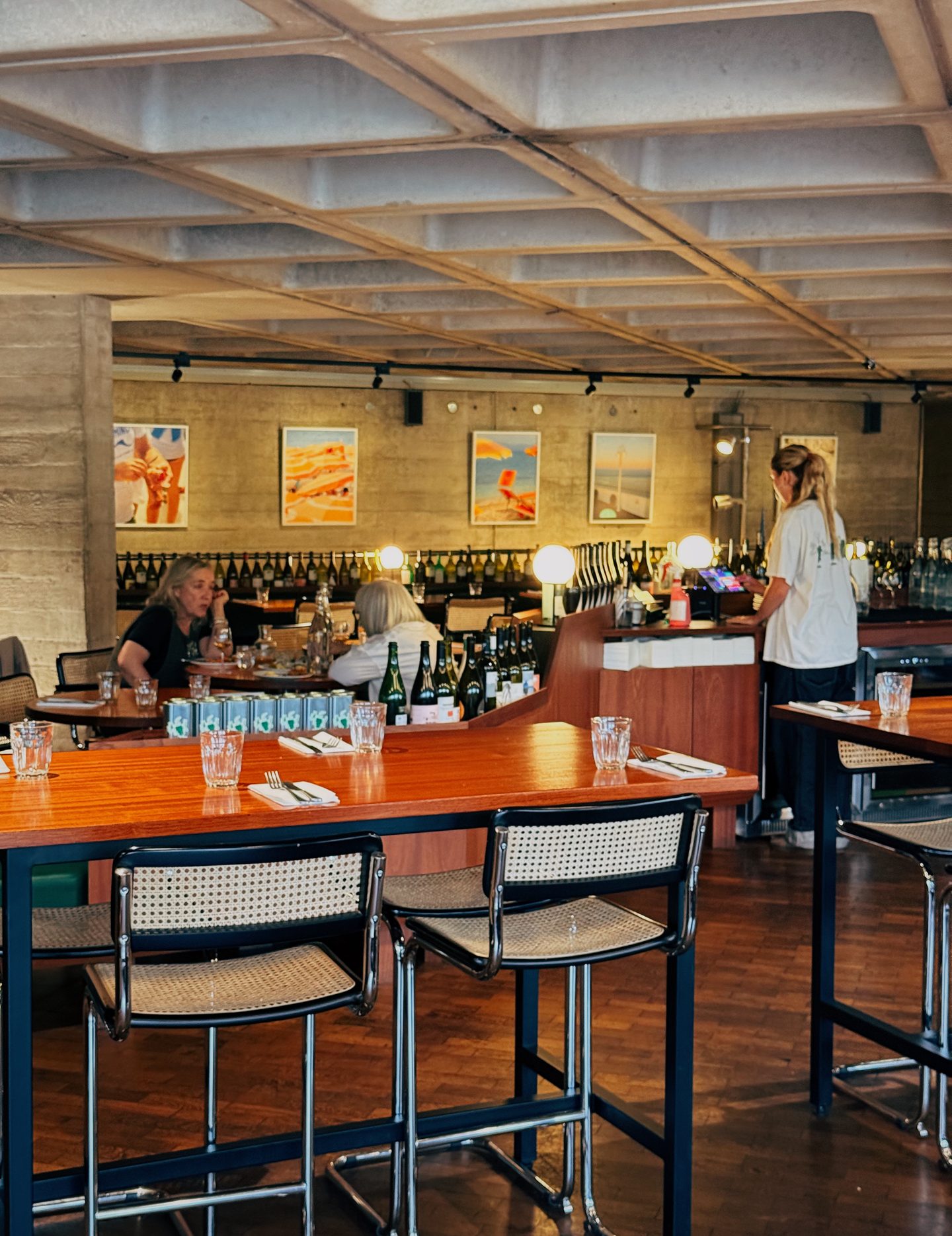 Interior of Forza Wine with long wooden tables cane chairs and bottles of wine lining the curved bar. Diners sit beneath colorful artwork in the warm modern dining room.