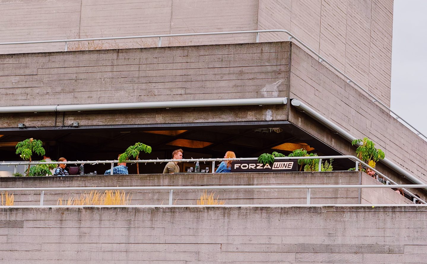 Diners sit among potted plants on the outdoor terrace at Forza Wine. The wine bar is tucked into the concrete architecture of the National Theatre on Londons South Bank.