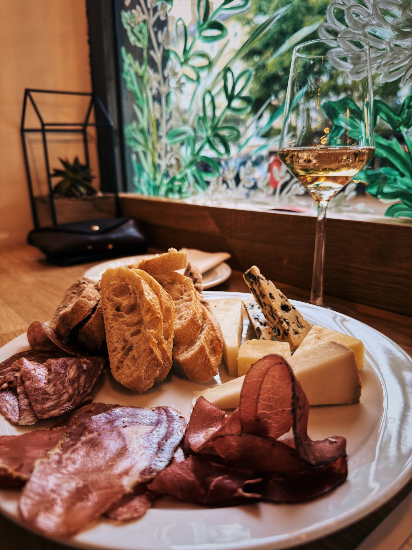Charcuterie and cheese board with sliced bread served beside a glass of wine at The Good Wine Shop. The table sits beside a decorative floral window looking out onto the street.