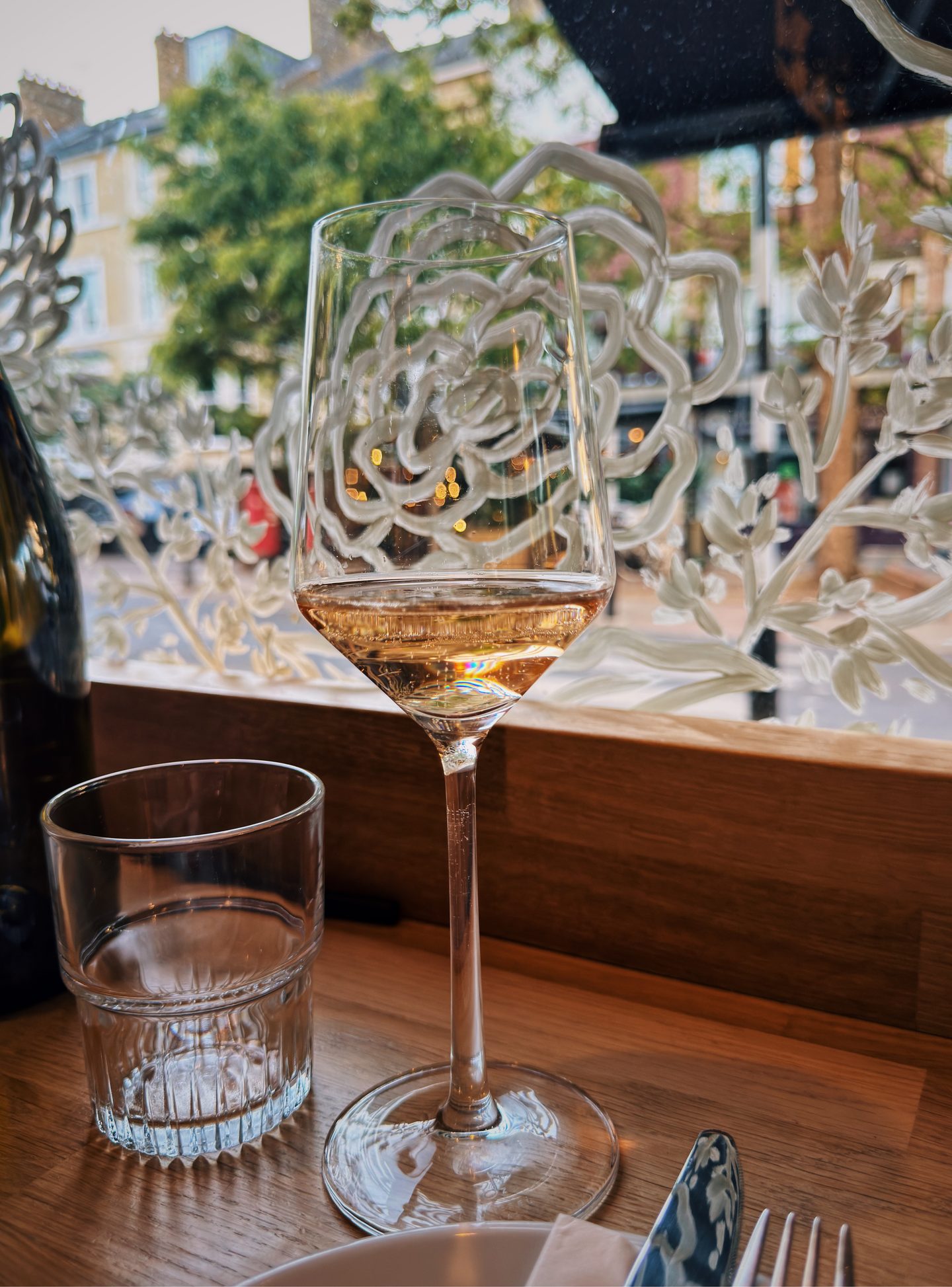Glass of pale wine and a water glass on a wooden table at The Good Wine Shop. An etched floral window frames a leafy street scene outside.
