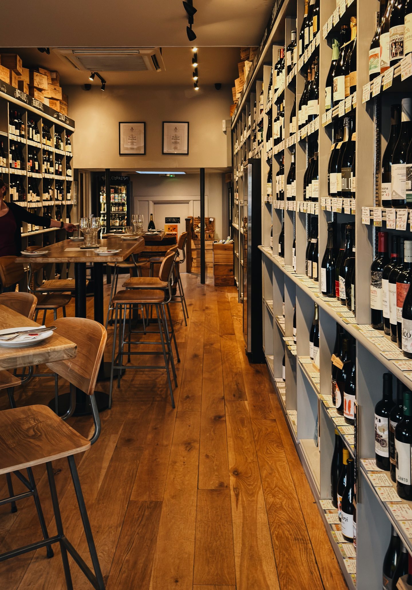 Interior of The Good Wine Shop with floor to ceiling shelves packed with wine bottles and handwritten labels. Wooden tables and bar stools run through the narrow warmly lit wine bar and shop.