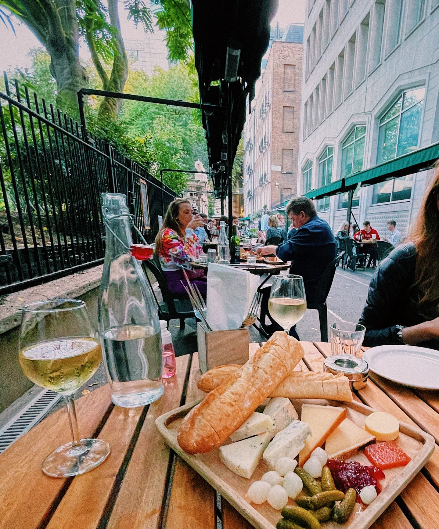 Cheese board with crusty baguettes pickles chutney and several cheeses beside glasses of white wine on the outdoor terrace at Gordons Wine Bar in central London. Diners fill the narrow terrace beside leafy greenery and surrounding city buildings.