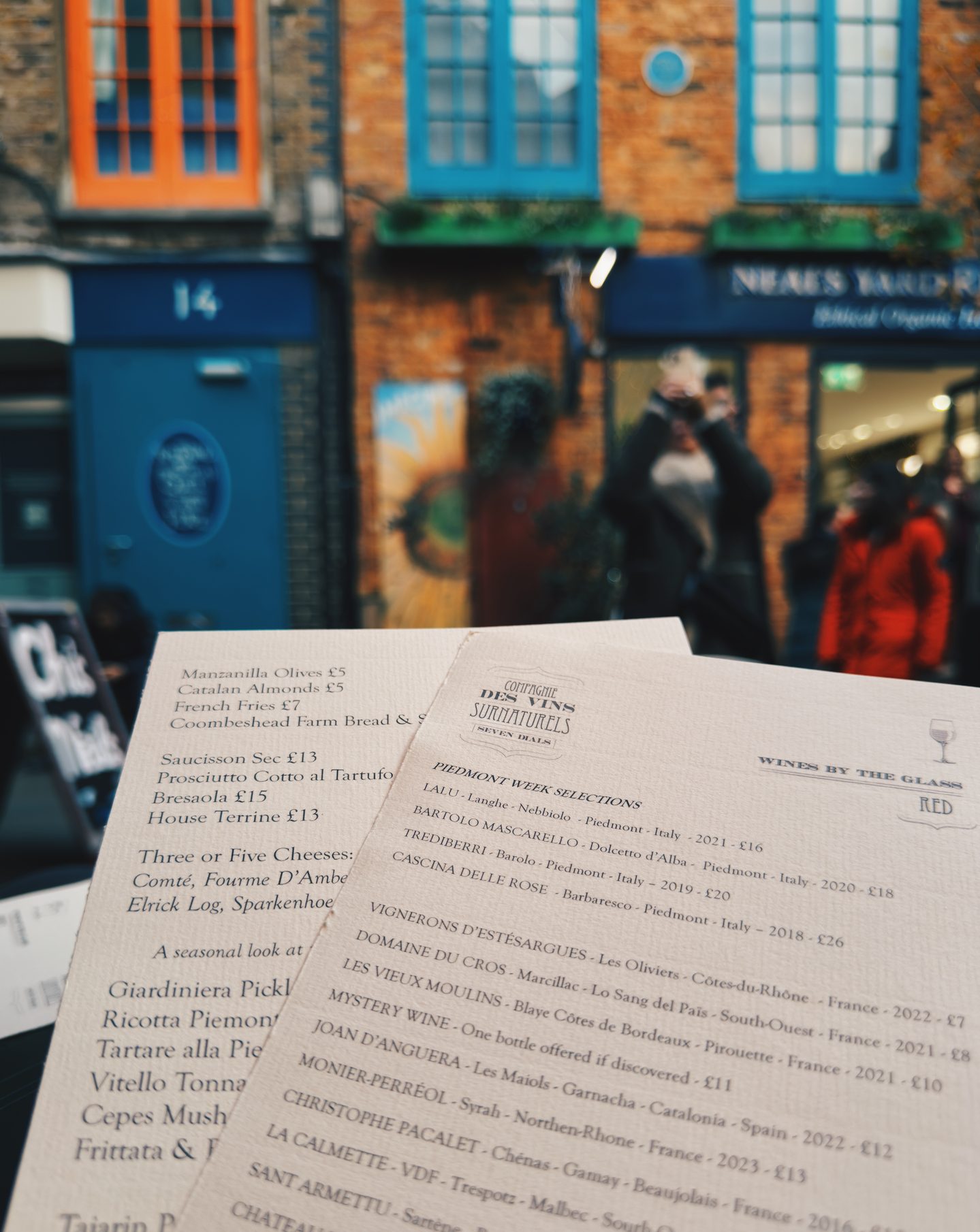 Wine and food menus at La Compagnie in colorful Neals Yard in Covent Garden with bright blue and orange storefronts and people in the background. A vibrant setting for a stop at one of the best London wine bars.