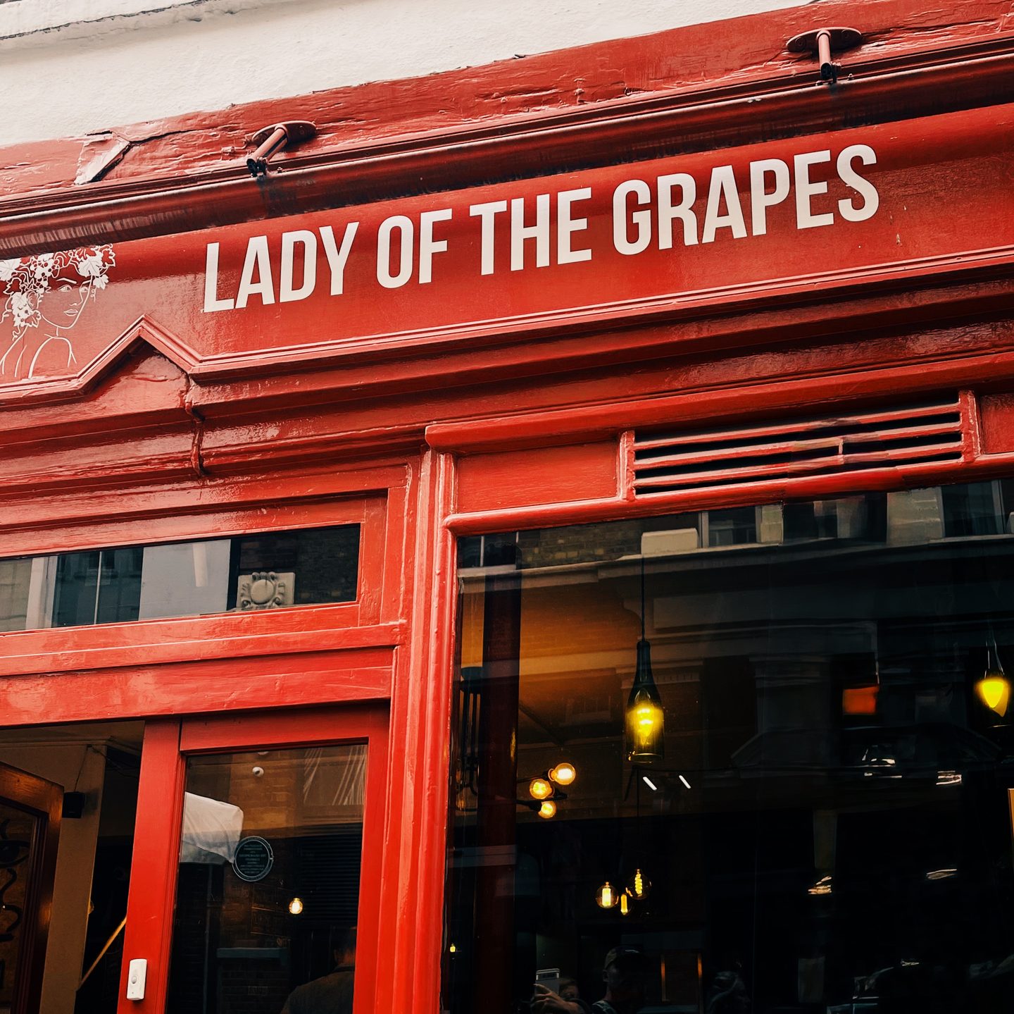Bright red exterior of Lady of the Grapes wine bar in central London with the name "LADY OF THE GRAPES" written in large white letters above the entrance. Warm pendant lights glow through the front window.