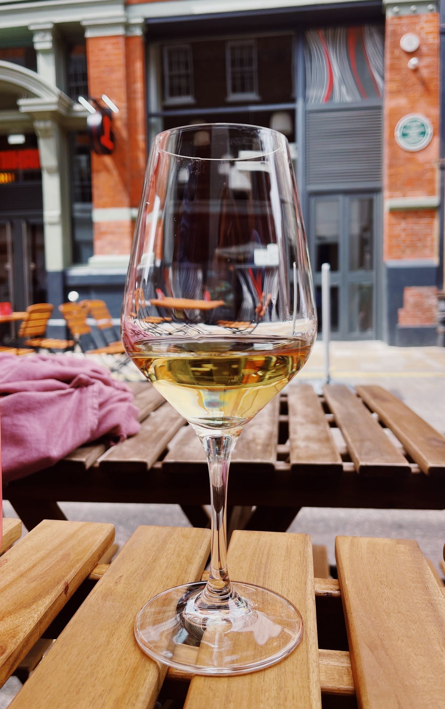 Glass of white wine on a wooden outdoor table at Lady of the Grapes wine bar in central London. The street side seating looks across to red brick buildings and more outdoor tables.
