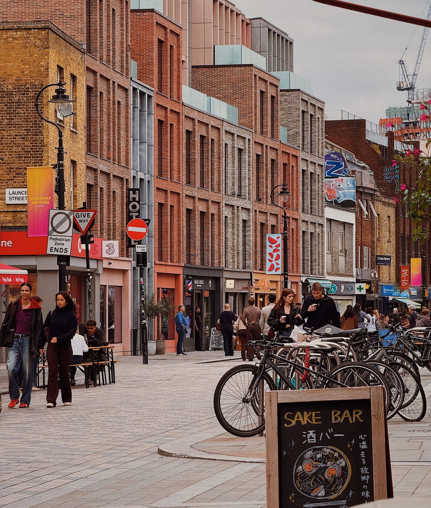 People walk along Lower Marsh in Waterloo past red brick buildings outdoor tables and rows of parked bicycles. A chalkboard advertising a sake bar sits in the foreground of the lively pedestrian street near Lower Wine Bar.