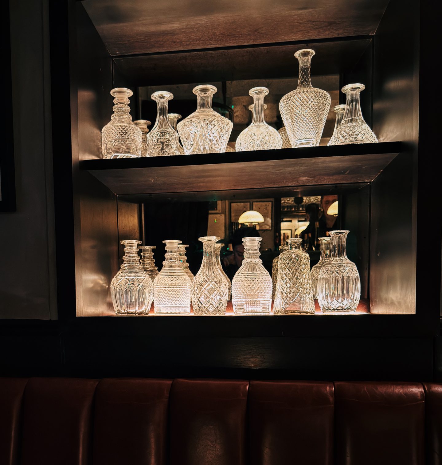 Illuminated crystal wine decanters displayed on dark shelves above a leather banquette at Noble Rot Mayfair in London. The glowing glassware adds to the intimate atmospheric feel of this celebrated London wine bar.