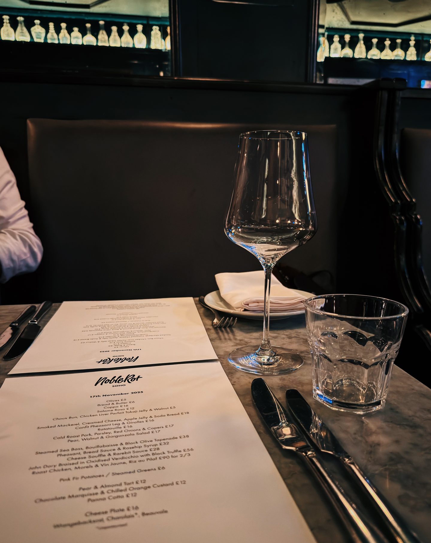 Table set for dining at Noble Rot Soho with an empty wine glass water glass cutlery and printed menus on a marble tabletop. Dark banquette seating and illuminated crystal decanters create a moody elegant setting for wine and dinner.