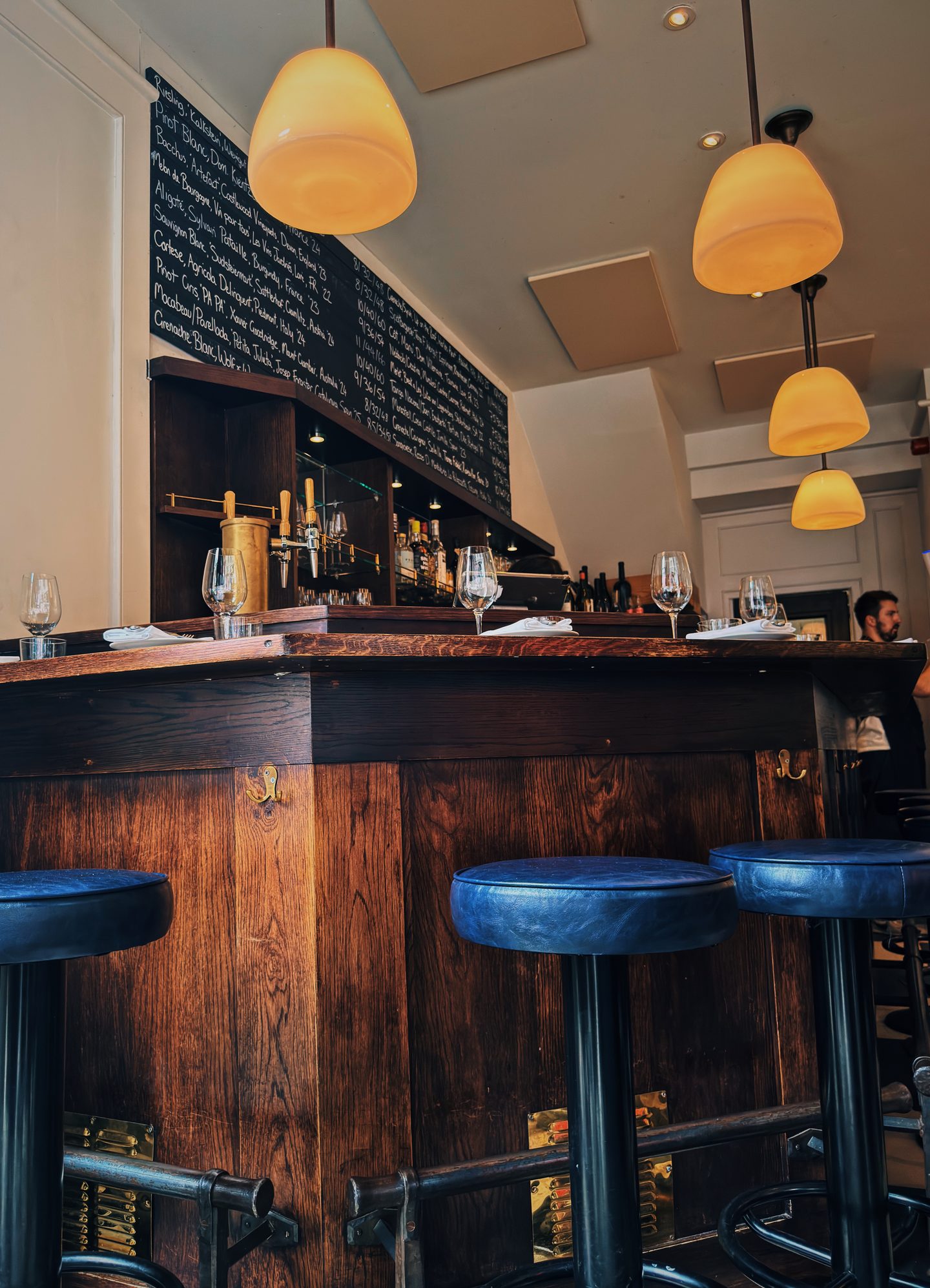 Interior of The 10 Cases in Covent Garden with a dark wooden bar blue stools warm pendant lights and a large handwritten wine list covering the wall. Wine glasses are set along the bar ready for guests.