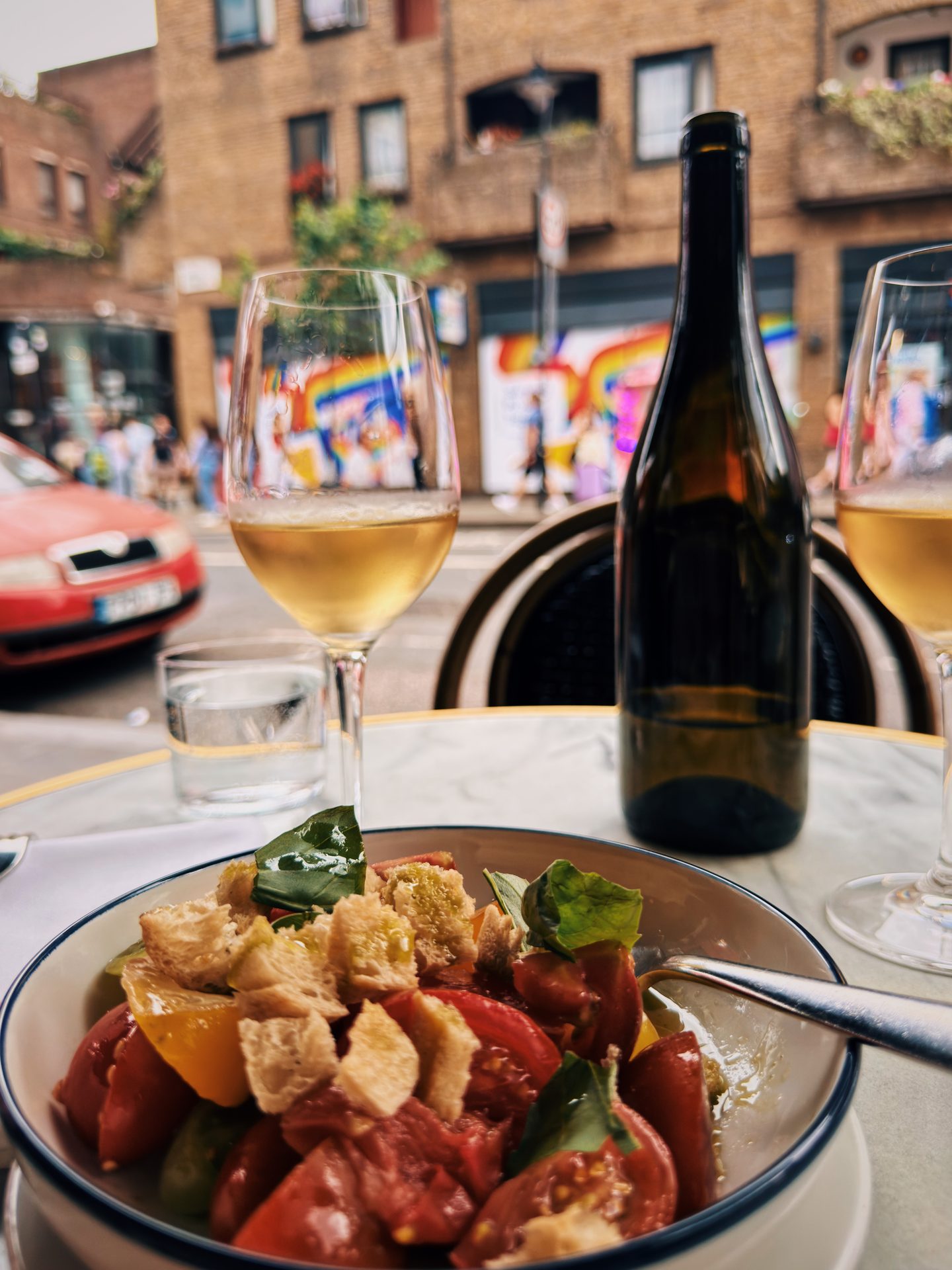 Pangrattato salad topped with crunchy croutons and fresh basil beside glasses of wine and a bottle at an outdoor table at The 10 Cases in Covent Garden. Colorful street art and brick buildings provide a lively London backdrop.