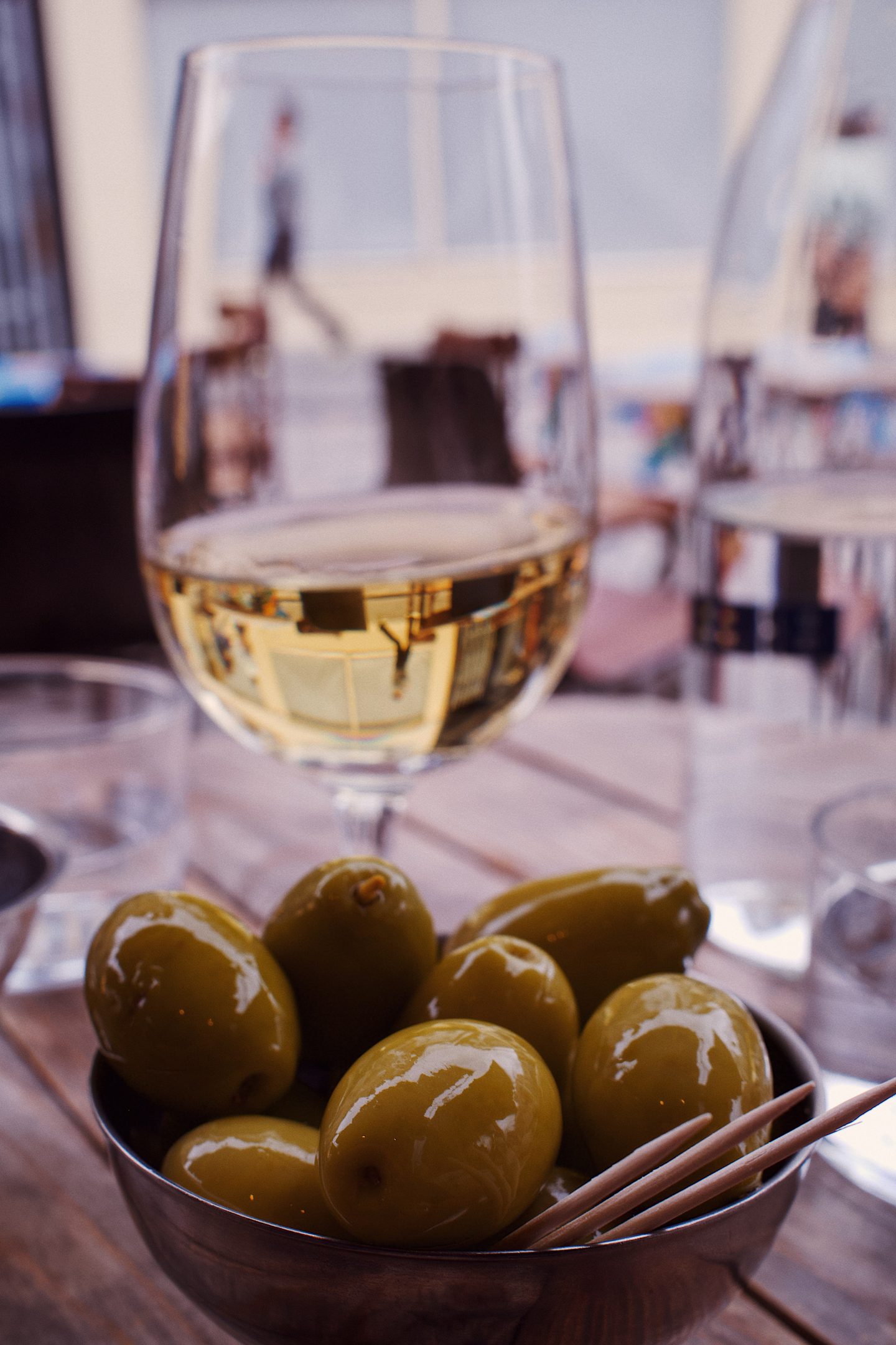 Glossy green olives served in a small metal bowl with cocktail sticks at Lower Wine Bar in Waterloo. A glass of white wine sits behind the bar snack.